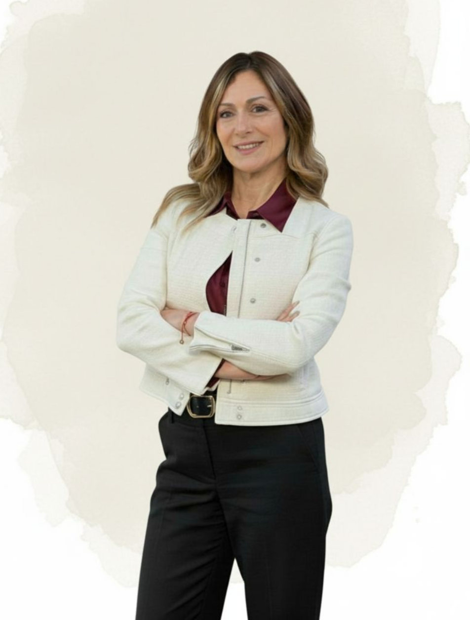 A woman with shoulder-length wavy hair, wearing a white jacket over a maroon blouse, standing with arms crossed in front of a plain light background.