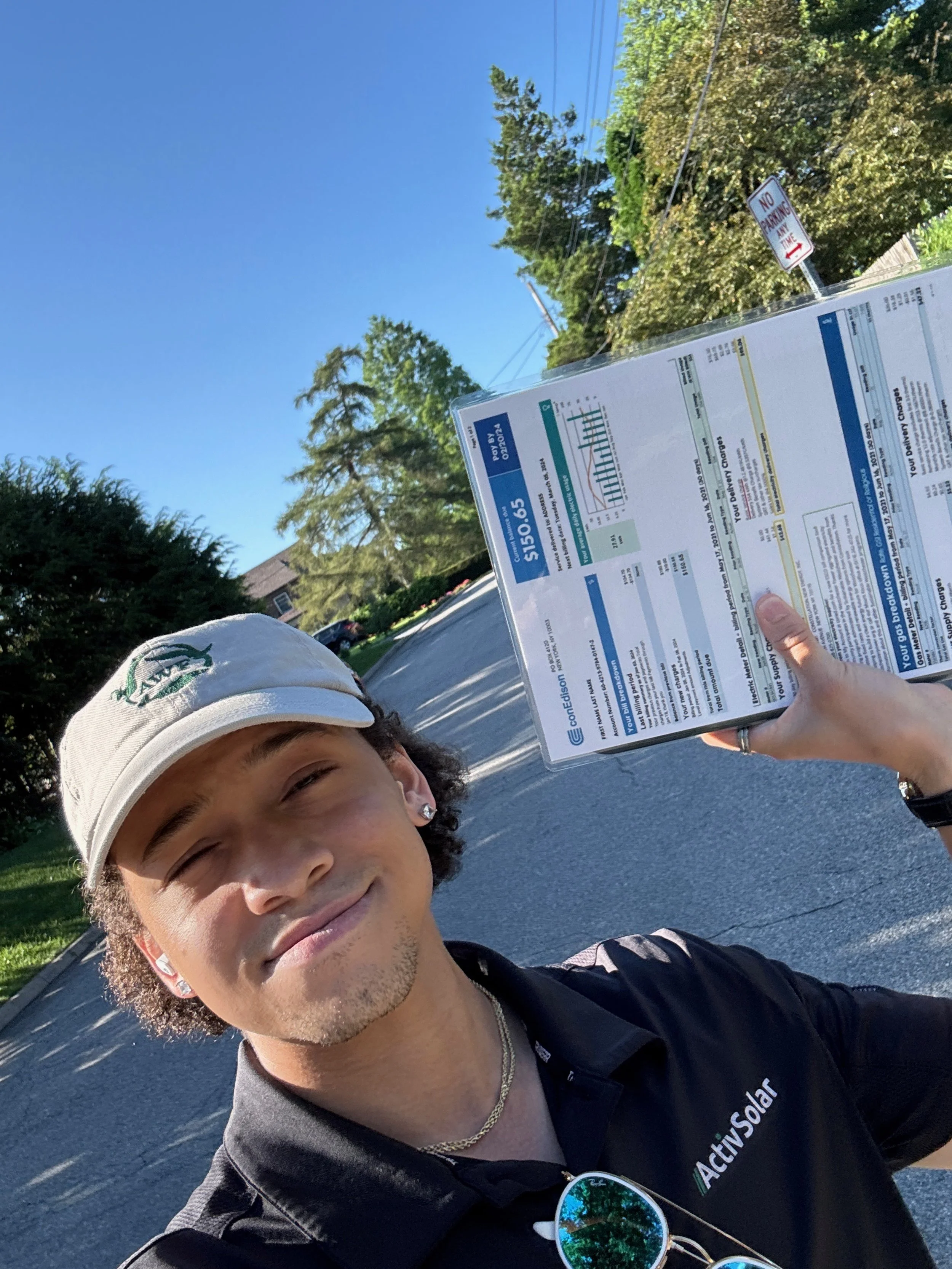A young man wearing a beige cap, earrings, and a black collared shirt with 'Activ Solar' on it, is standing outdoors on a sunny day holding a document or pamphlet with charts and text, with trees and a clear blue sky in the background.