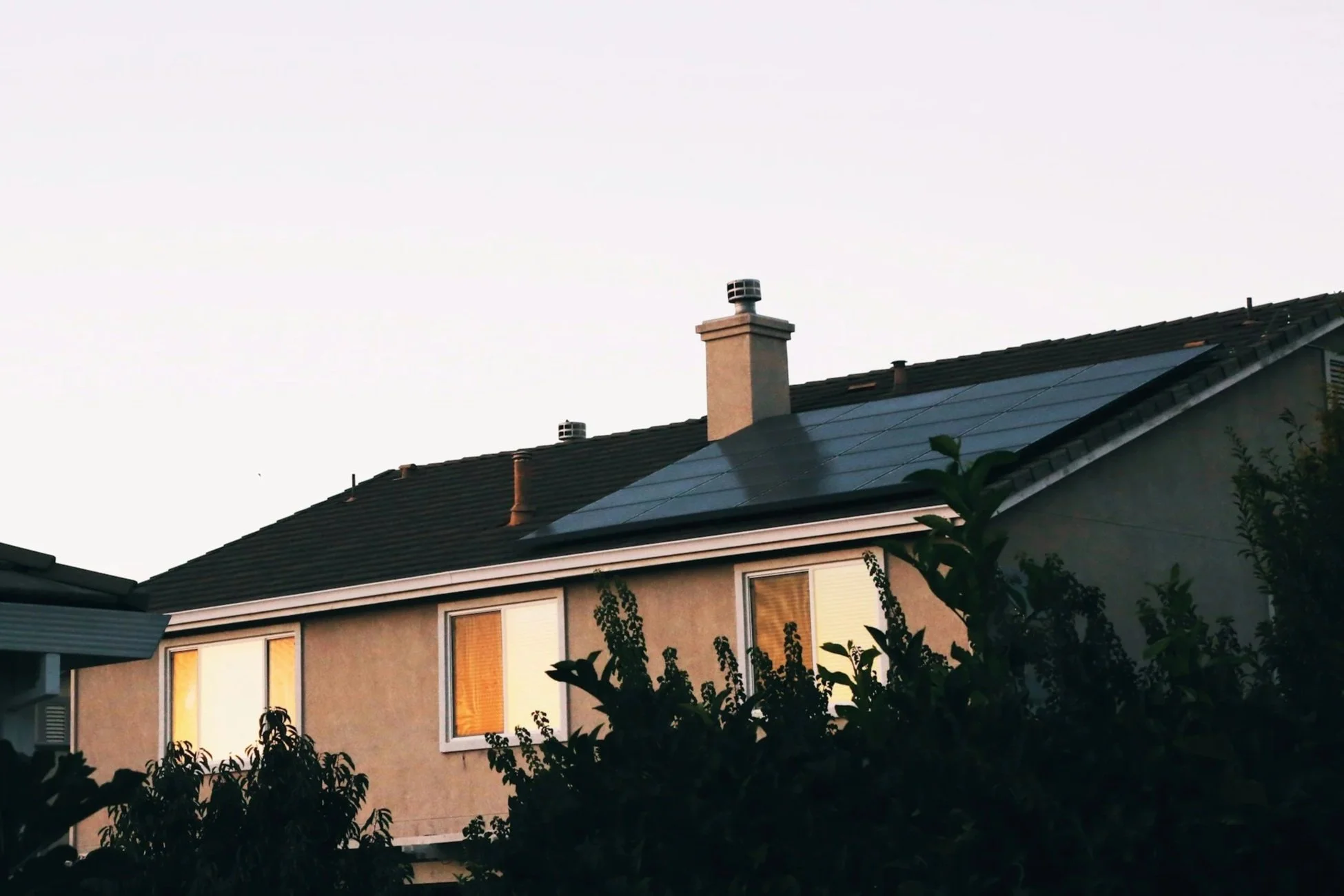A house with solar panels on the roof, illuminated windows at sunset, and greenery in front.