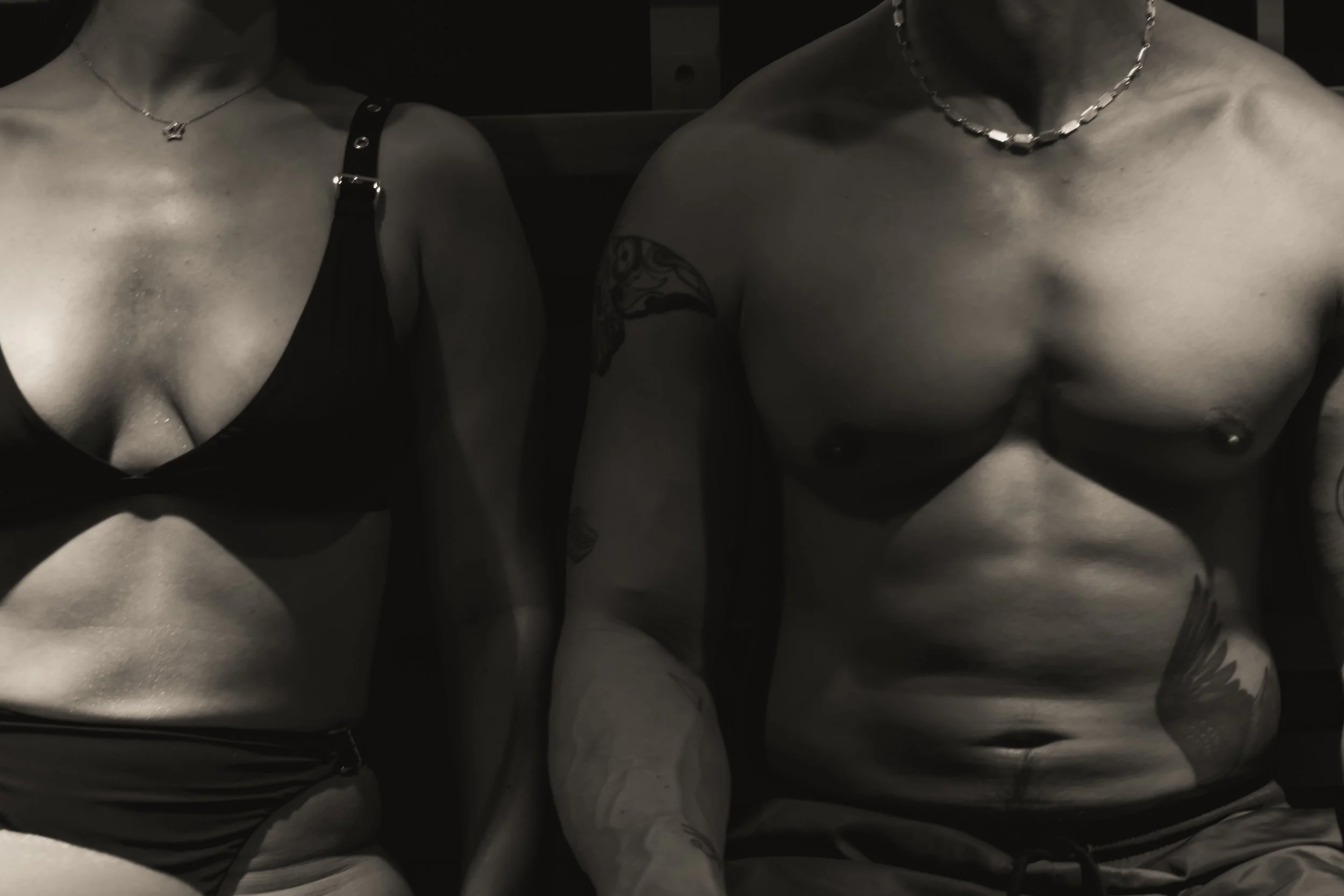 Black and white photo of a topless man and woman with tattoos, wearing necklaces, sitting side by side.