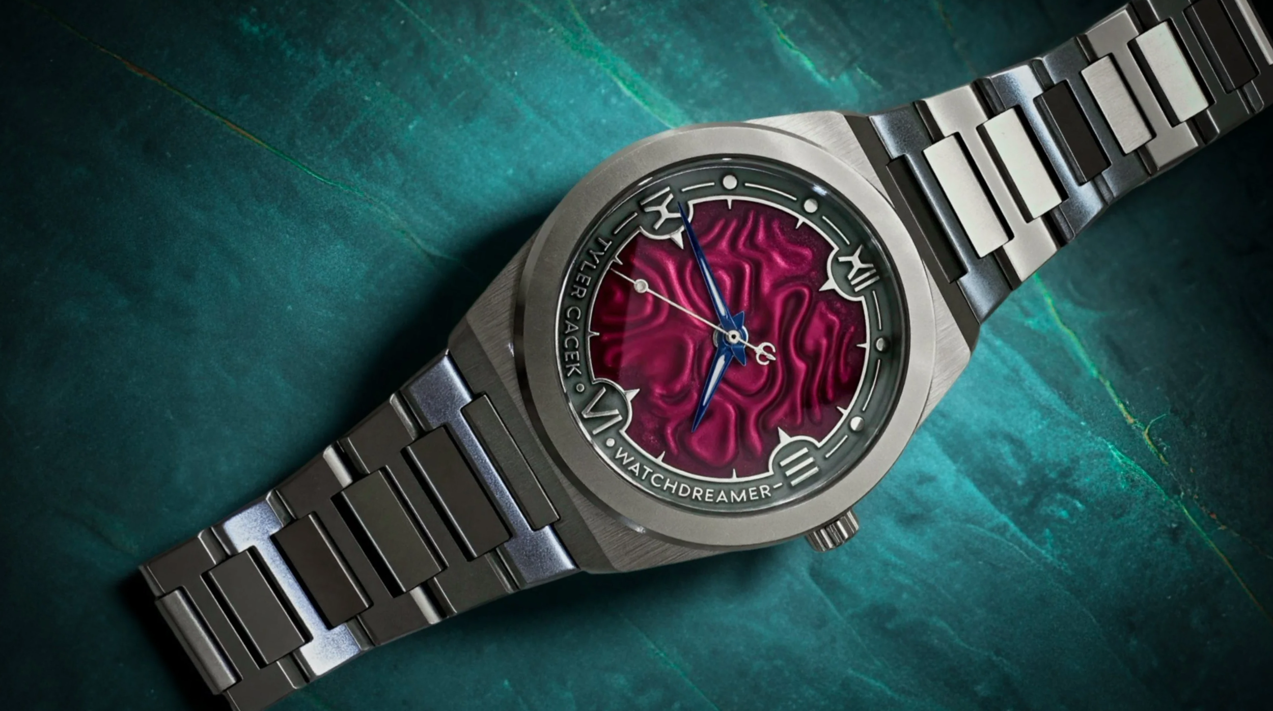 A luxury wristwatch with a metallic band, round face, and a vibrant pink textured dial. The watch hands are blue, with the hour and minute hand being ornate. The face displays Roman numerals at 3, 6, 9, and 12 o'clock positions, and the watch brand "V. WATCHDREAMER" is visible on the dial. The watch is placed on a dark green textured surface.