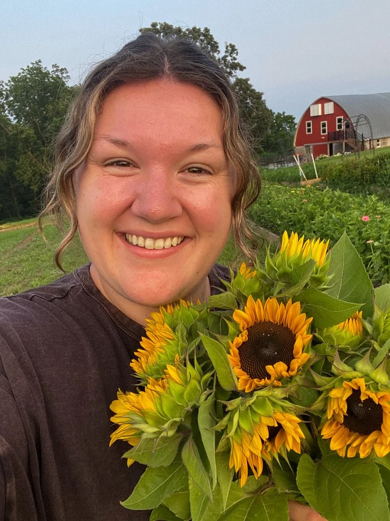A woman smiling and holding a bouquet of yellow sunflowers with green leaves, standing outdoors on a farm with a red barn and green fields in the background.