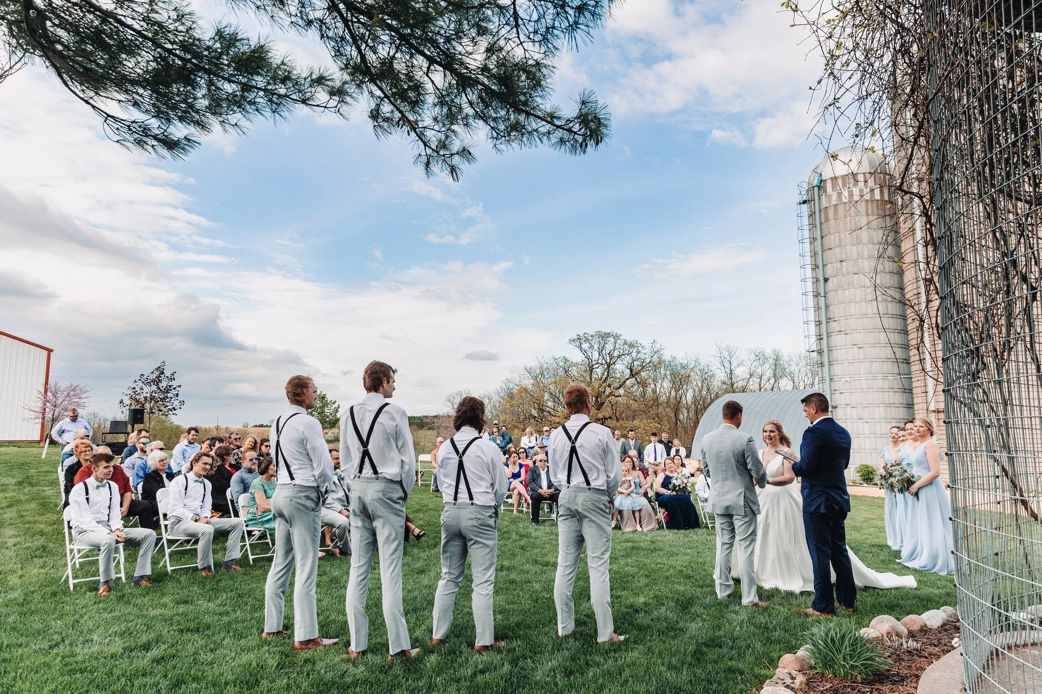 Outdoor wedding ceremony with the couple, officiant, and wedding party on lush green grass, surrounded by seated guests, with a farm silo in the background under a partly cloudy sky.