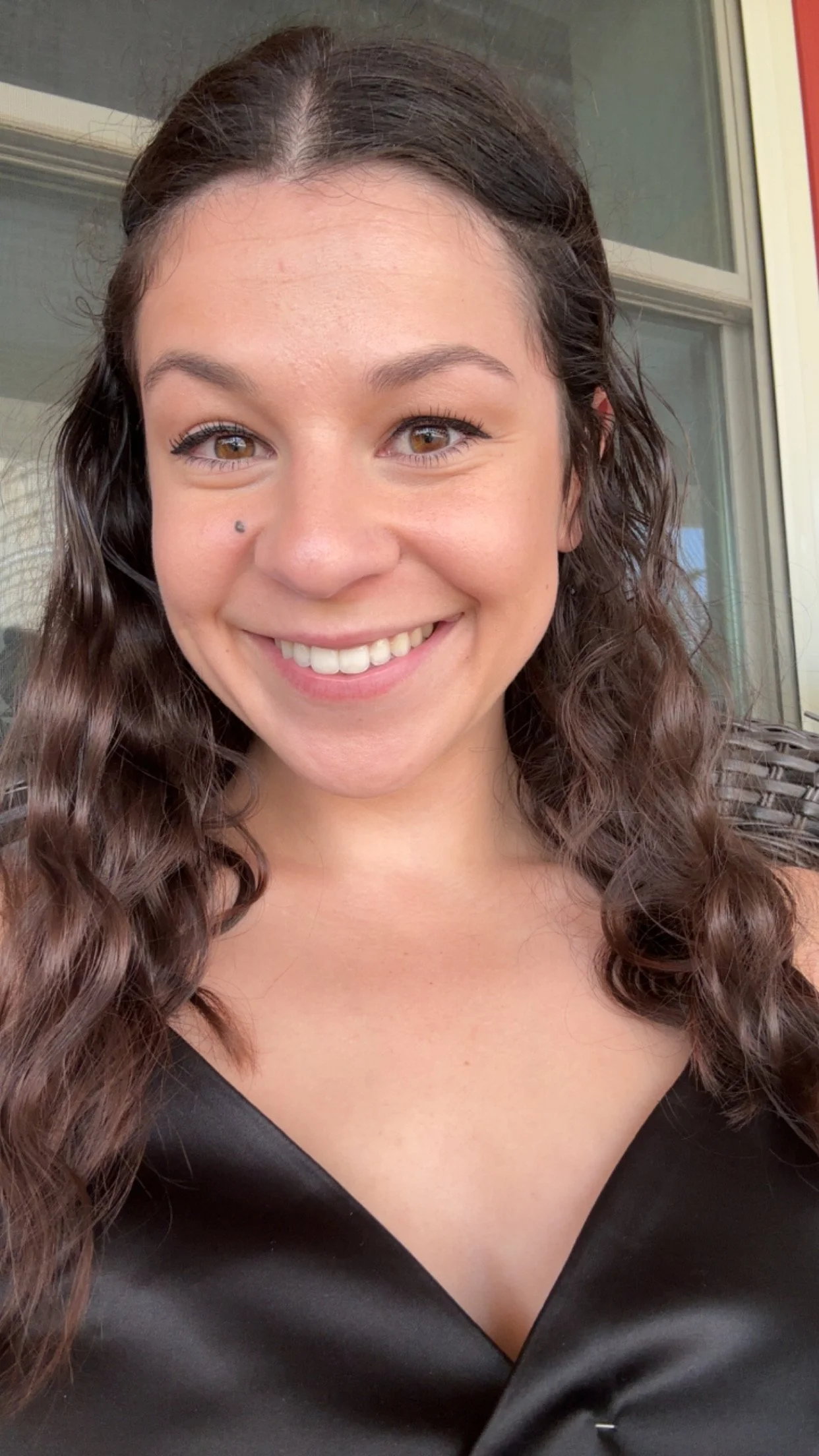 A woman with long, curly brown hair, brown eyes, and a small mole on her left cheek, smiling at the camera.