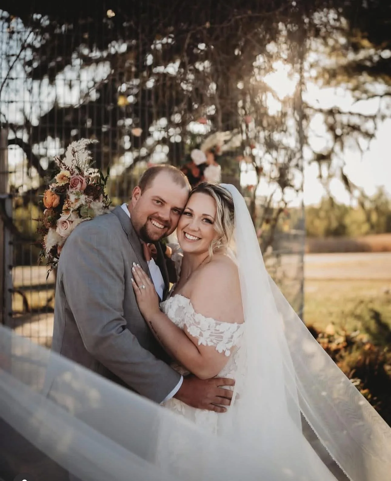 A newlywed couple smiling and embracing outdoors during sunset. The groom is wearing a gray suit and the bride is in a white wedding dress with off-shoulder lace sleeves and a veil. There is a floral bouquet behind them and trees in the background.