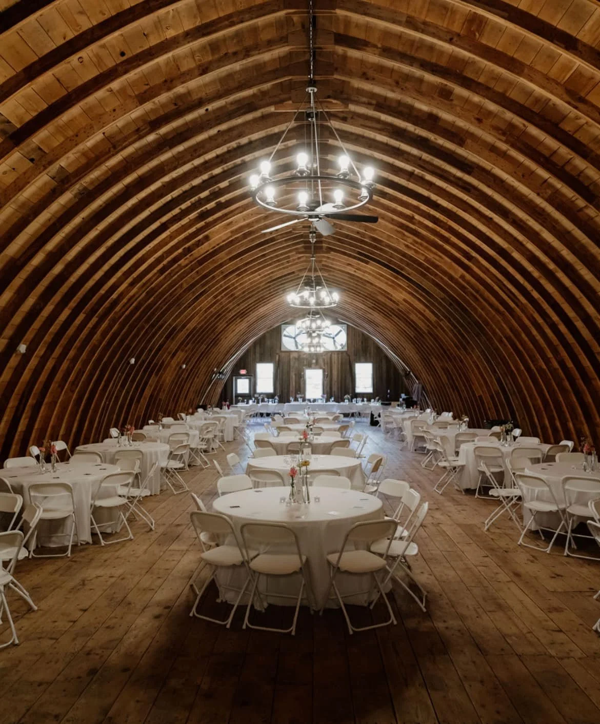 An arched wooden ceiling with chandeliers, set up for a banquet with round tables covered in white tablecloths and arranged with chairs, small floral centerpieces, and glassware.