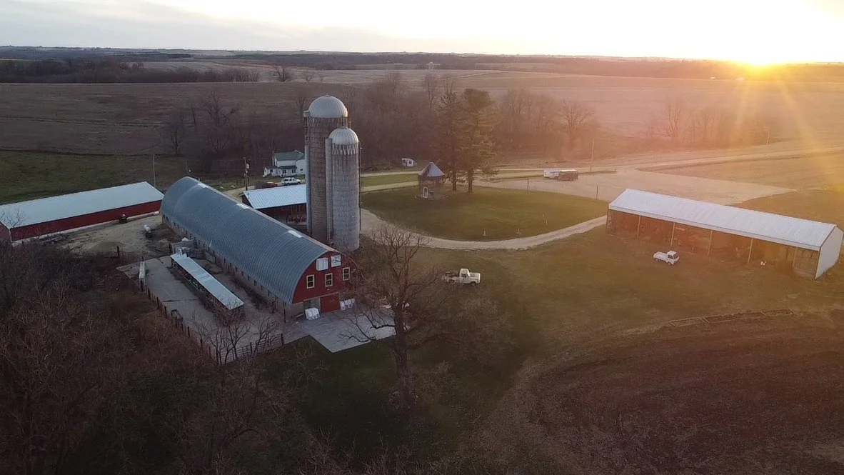 An aerial view of a farm at sunset, featuring a red barn with a curved metal roof, a large concrete silo, a smaller wooden silo, and several farm trucks and vehicles on grassy fields surrounded by trees and open land.