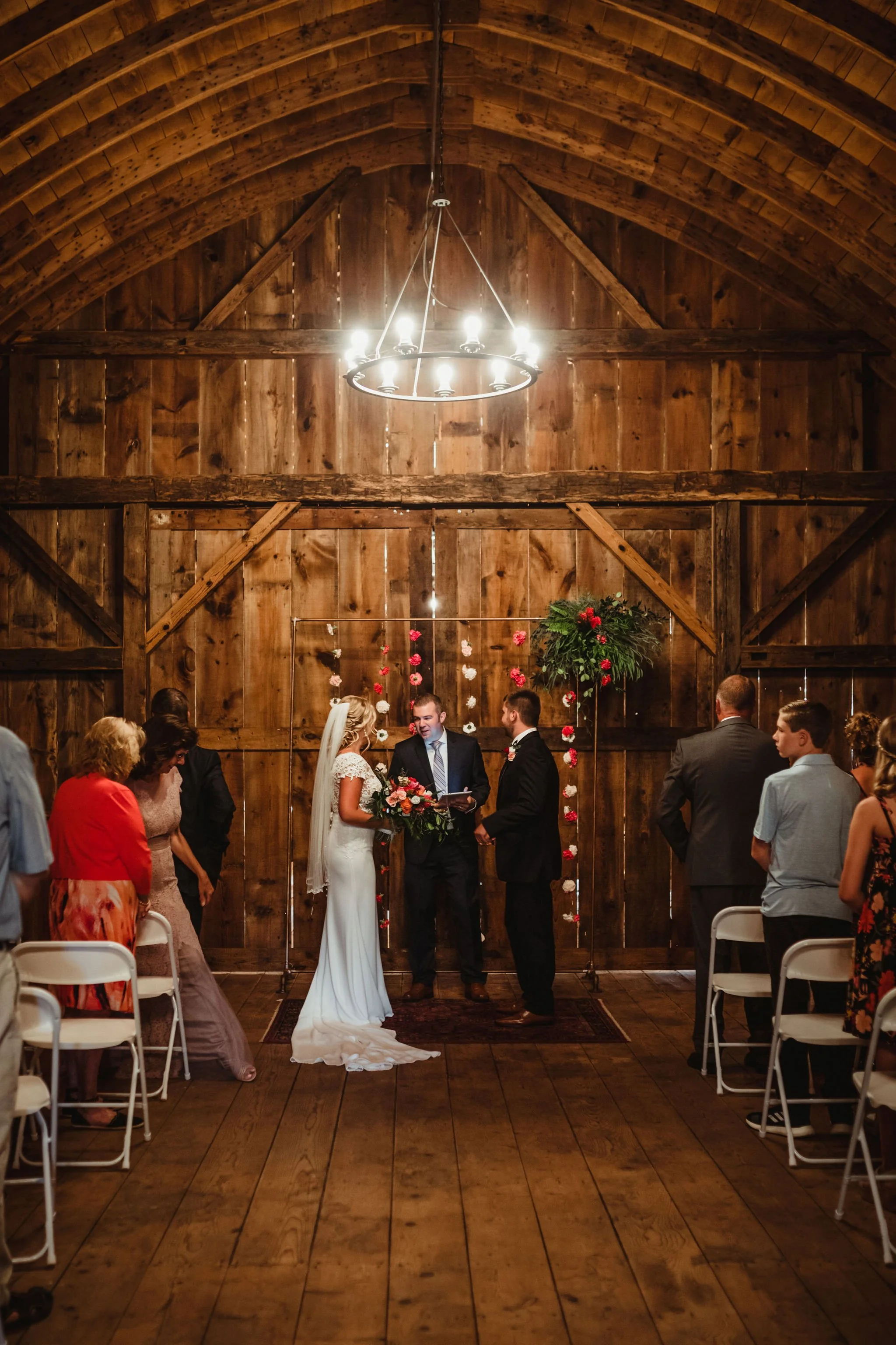 A couple gets married in a rustic barn, standing before a small audience of family and friends, with floral decorations and hanging greenery behind them.