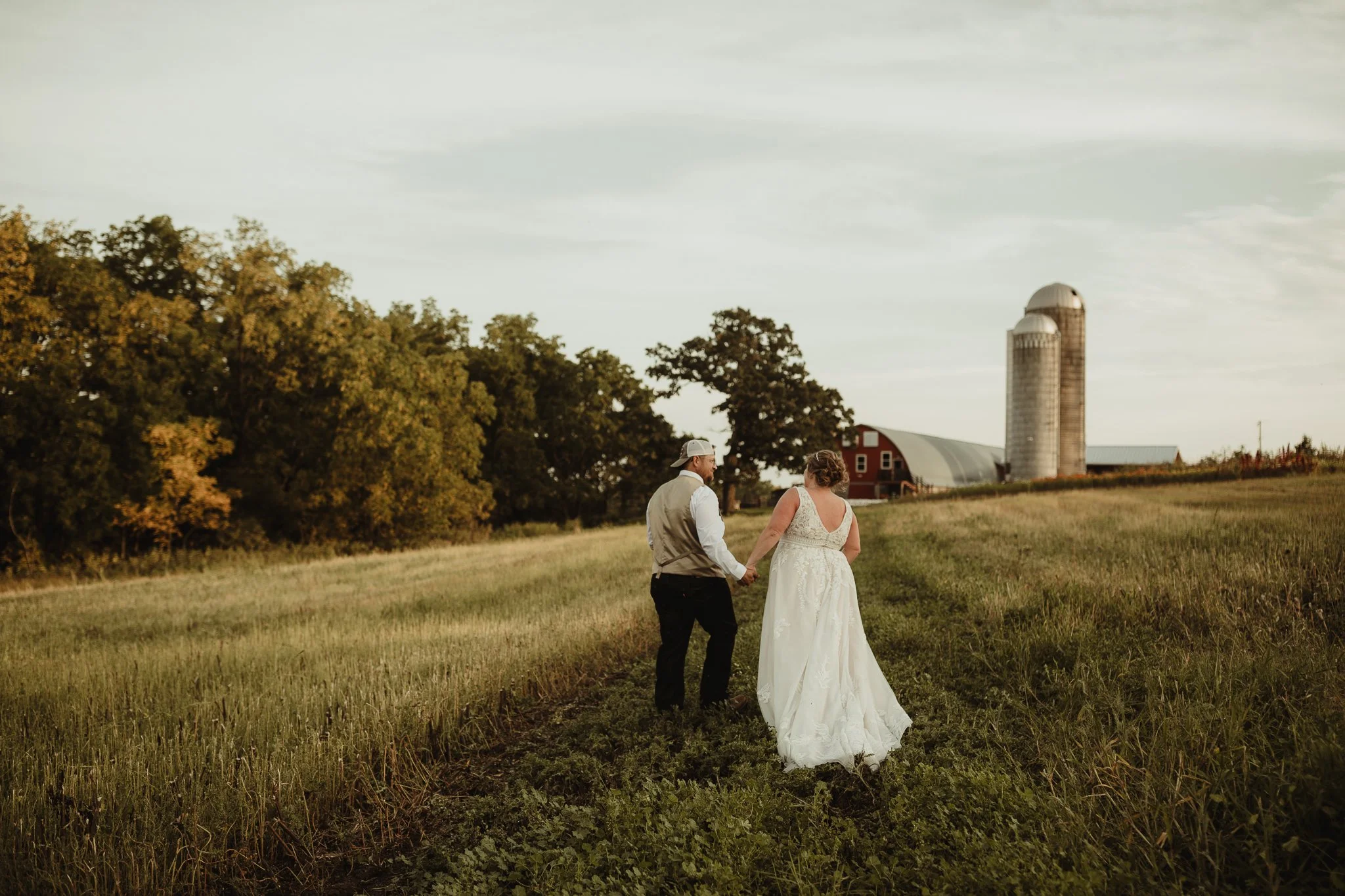 A bride and groom walking hand in hand through a grassy field at sunset, with a red barn and silos in the background.