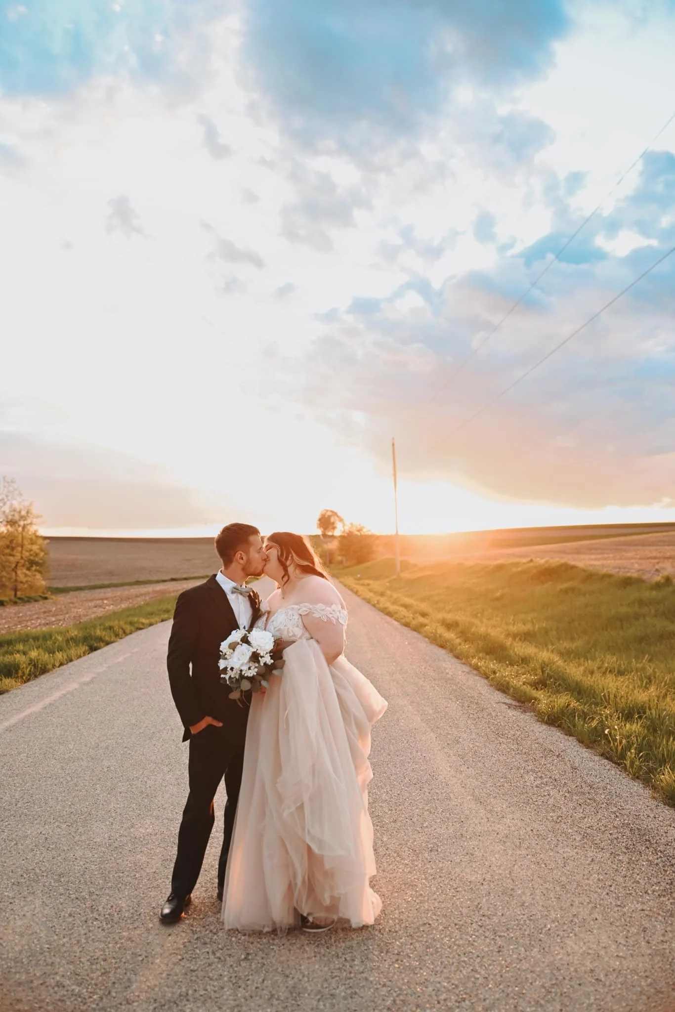 A bride and groom sharing a kiss on a rural road at sunset, with open fields and a partly cloudy sky in the background.