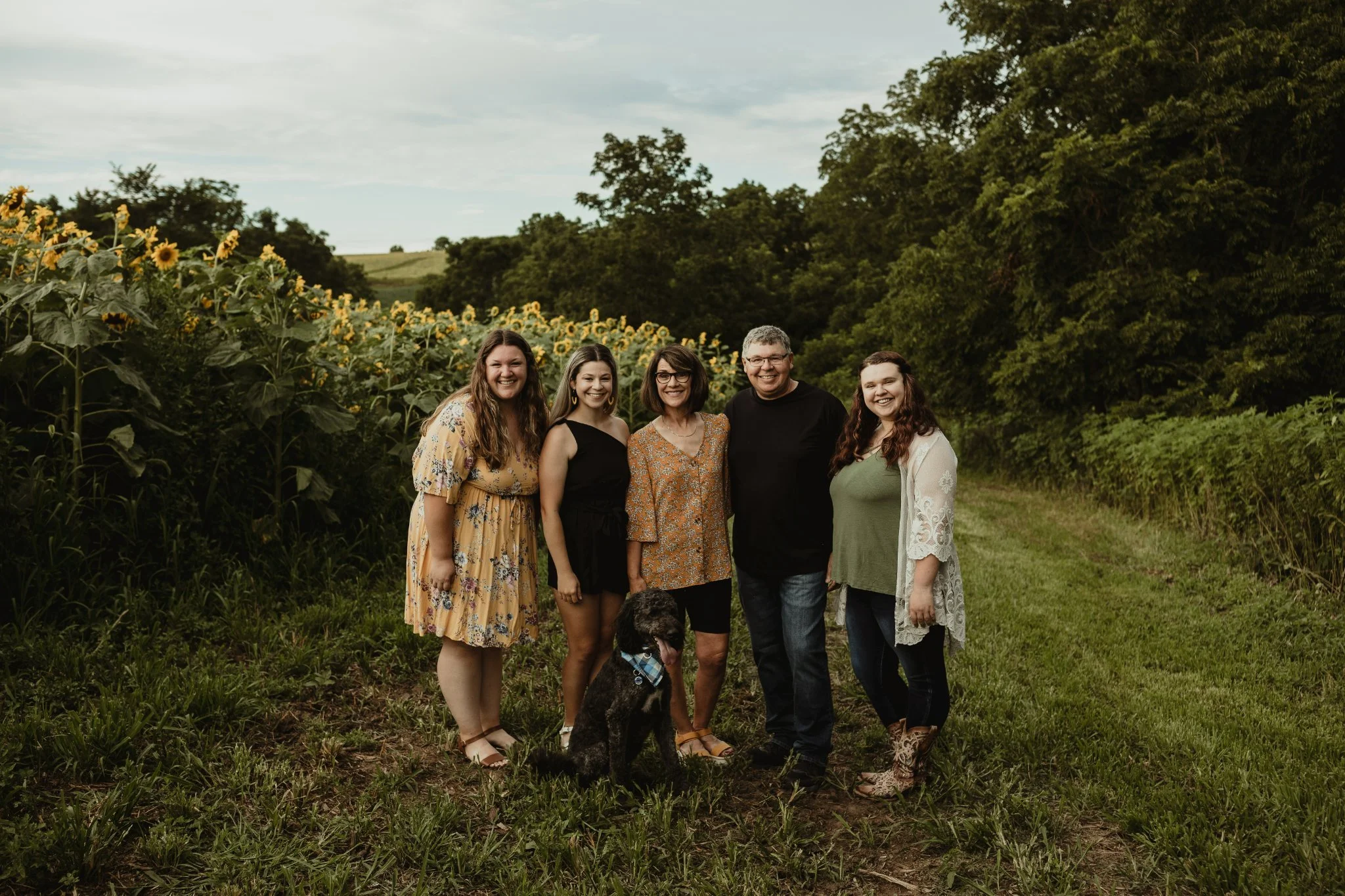 Family of five with a dog standing in a field with sunflower plants and green trees in the background.