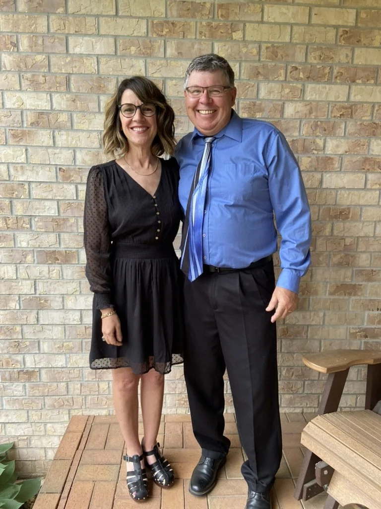 A man and woman standing side by side, smiling, in front of a brick wall. The woman is wearing glasses and a black dress with sheer long sleeves and black sandals. The man is dressed in a blue shirt, striped tie, and black pants.