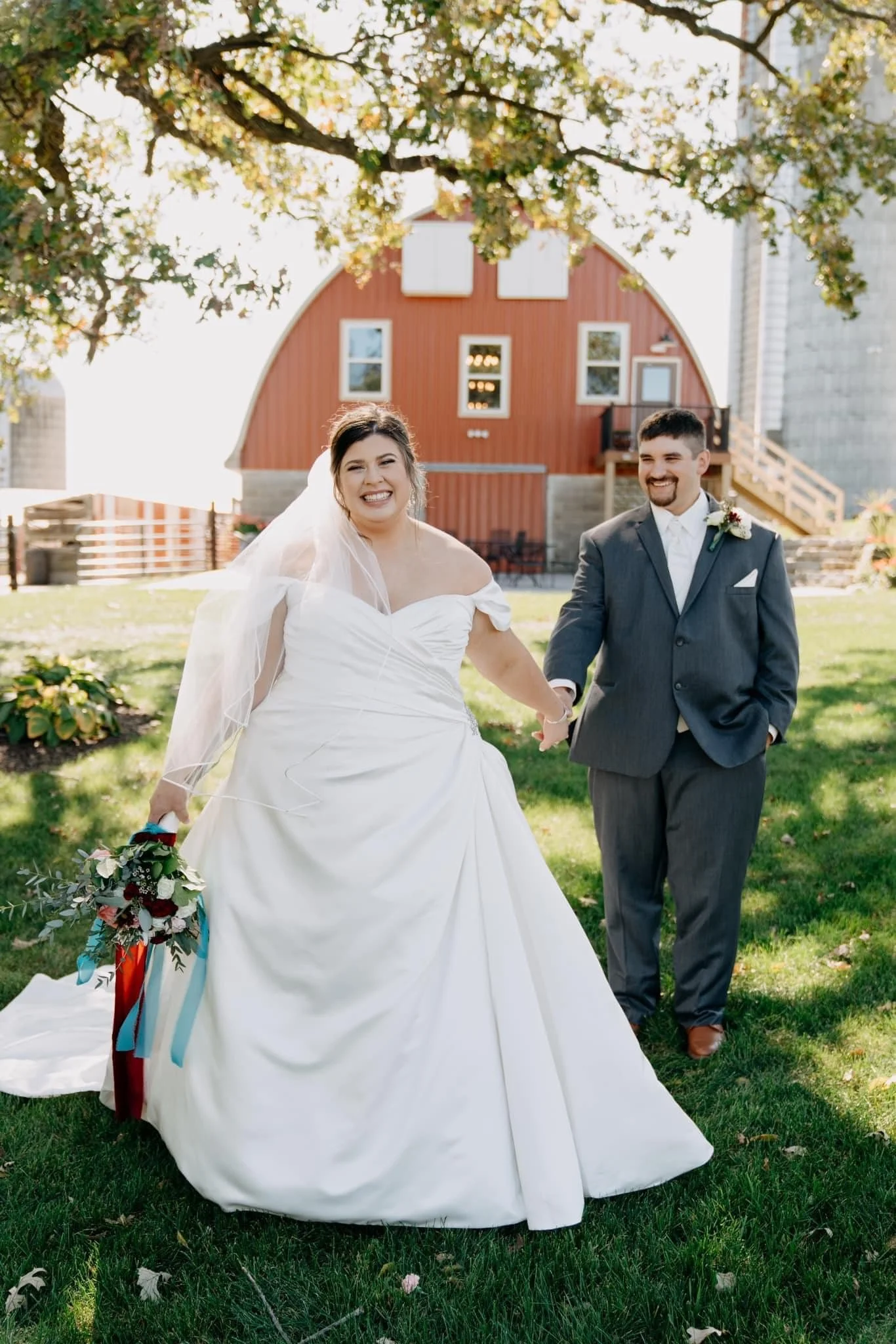 A bride and groom walking hand-in-hand in a grassy outdoor area with trees and a red barn in the background on a sunny day.