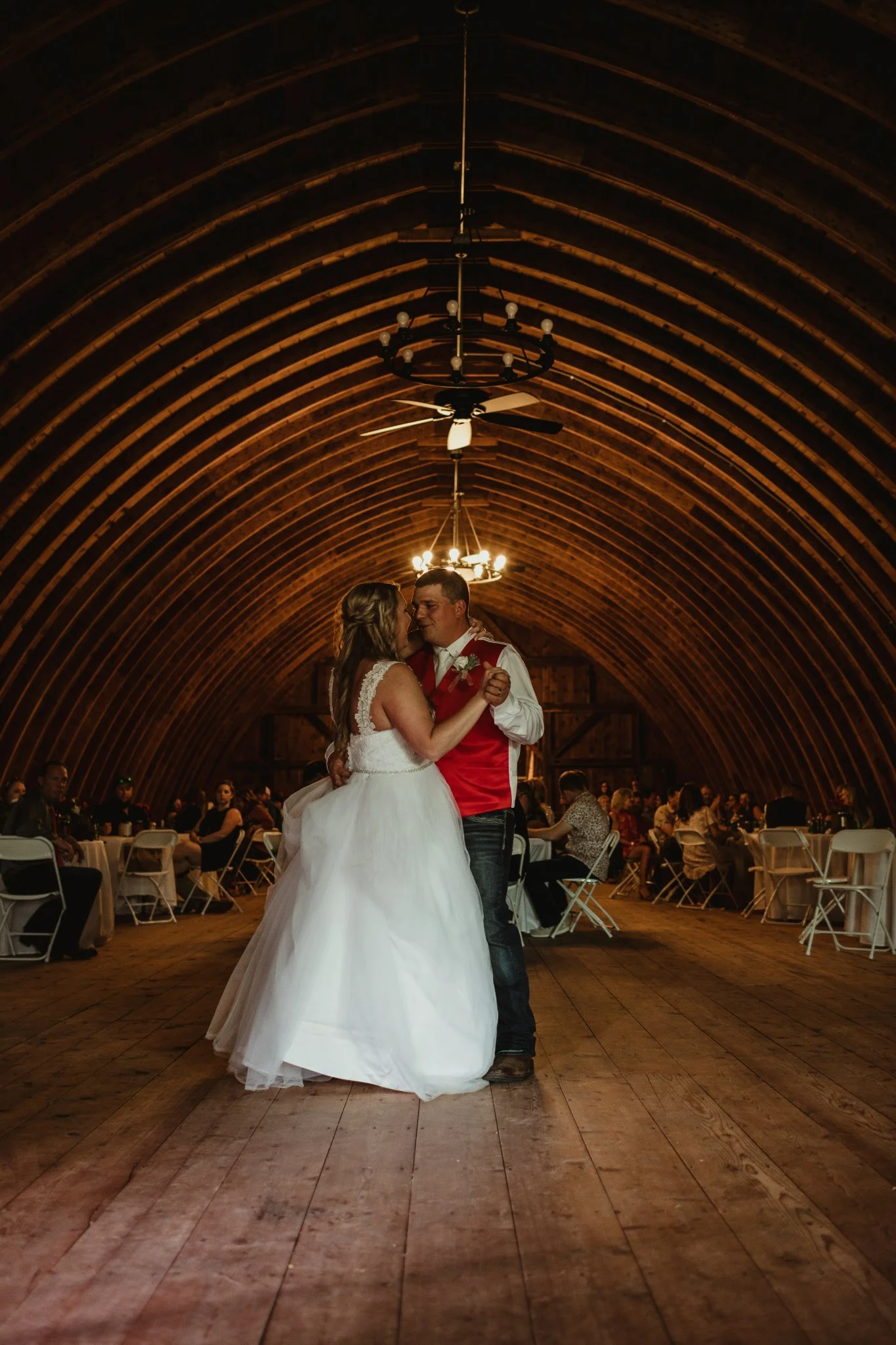 A bride and groom sharing a dance in a rustic barn with arched wooden ceiling and chandeliers