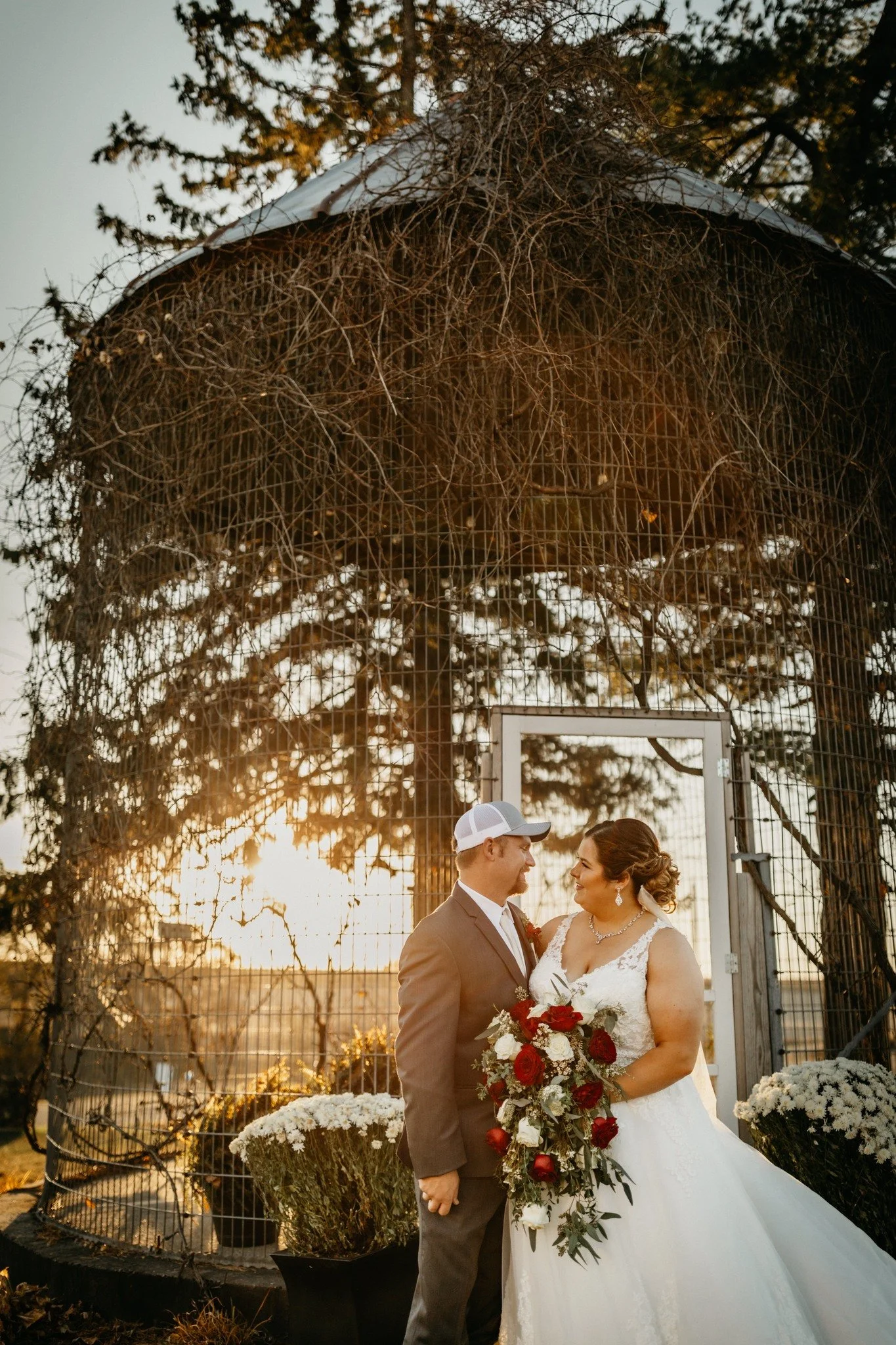 Couple in wedding attire standing close together outdoors during sunset, with a large wire structure behind them and flowers in the scene.