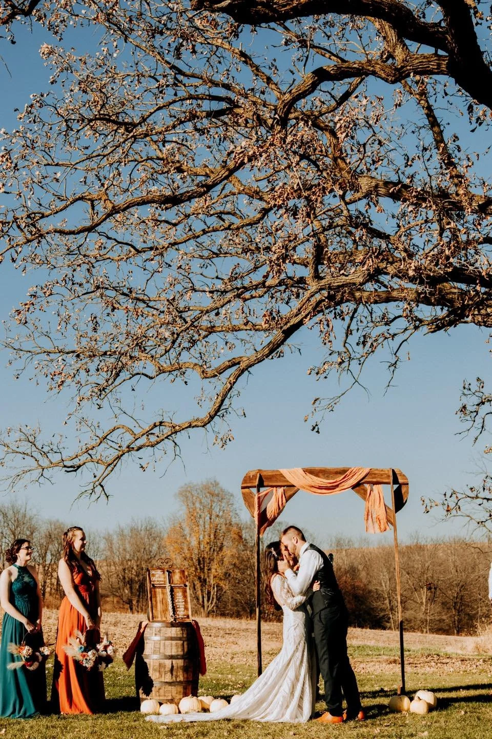 A wedding ceremony outdoors under a large tree with colorful dresses and a couple embracing and kissing.