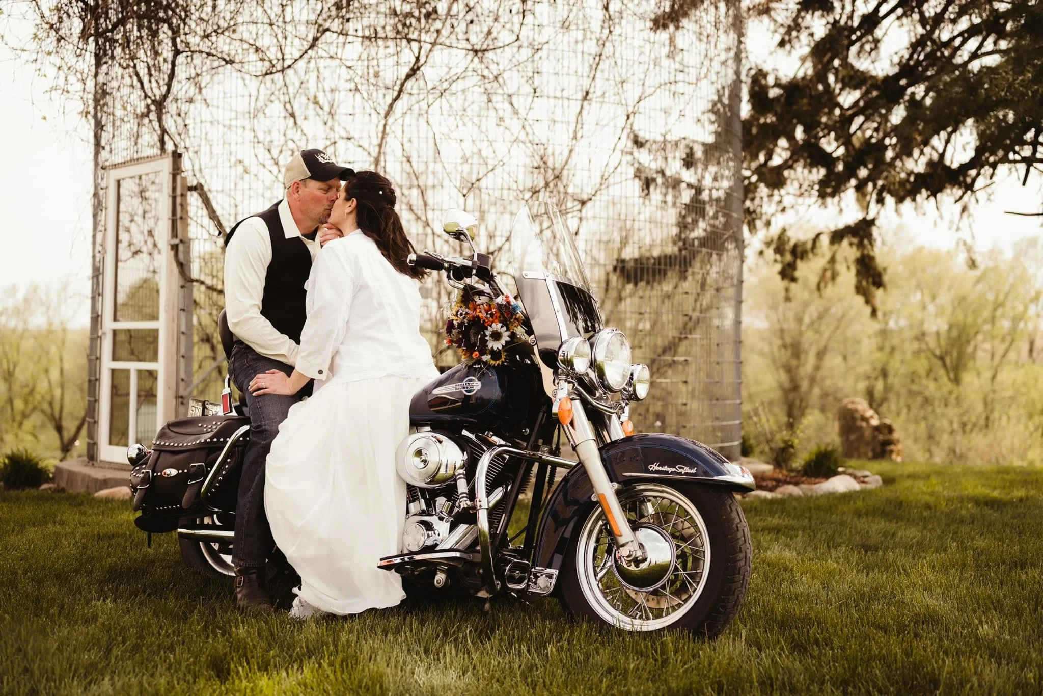 A man and woman share a kiss on a black Harley-Davidson motorcycle outdoors, with trees and a wire fence in the background.