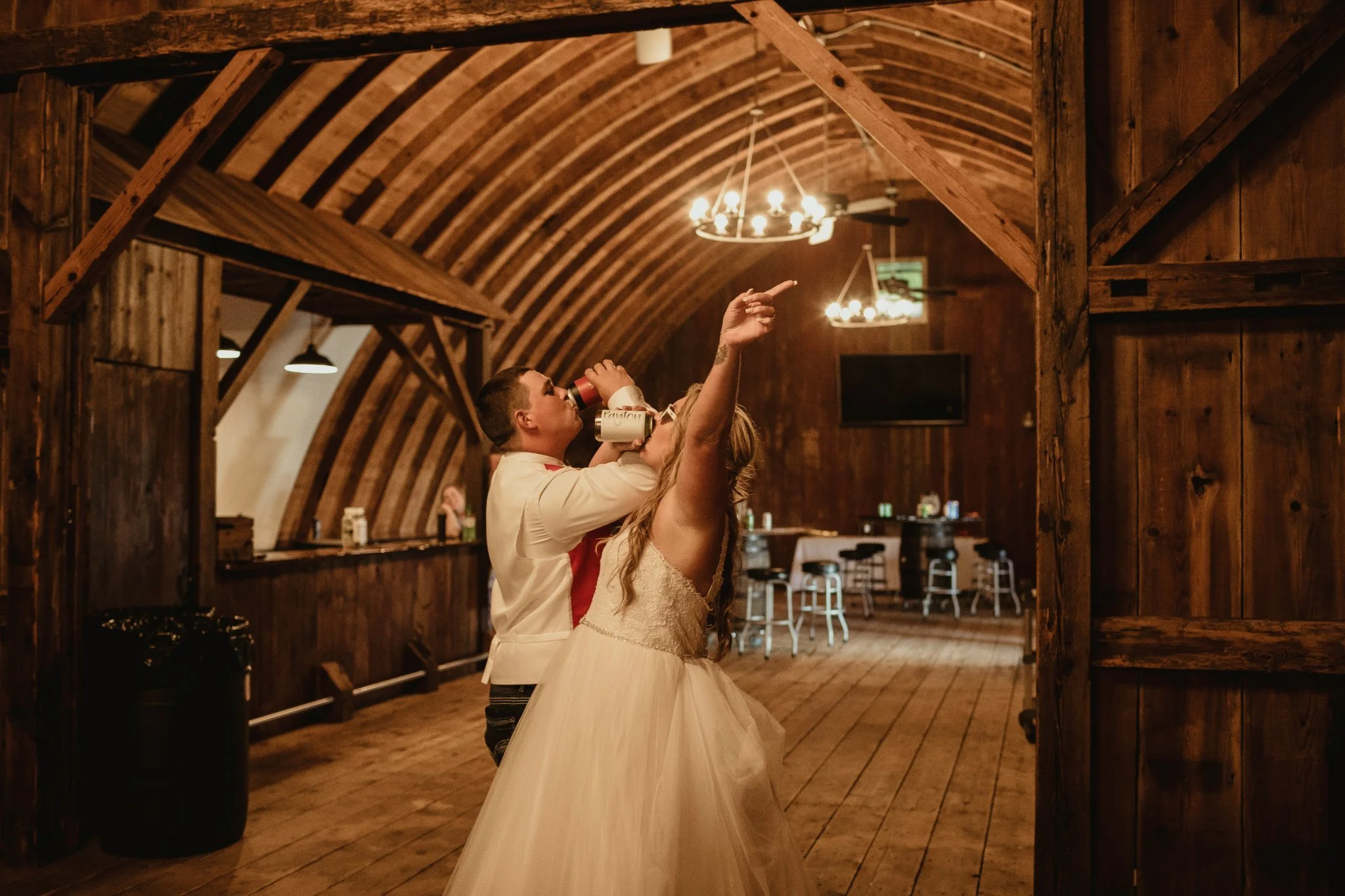 A couple dancing at a wedding reception in a wooden barn, with chandeliers hanging from the arched ceiling, and the man drinking from a bottle while pointing upwards.