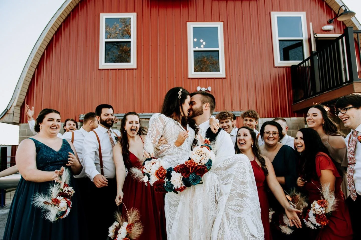A newlywed couple in wedding attire sharing a kiss, surrounded by smiling friends holding bouquets, outside a red barn building.