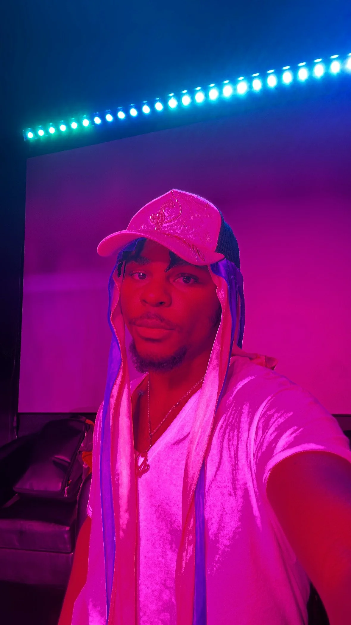 A young man taking a selfie in a room illuminated with pink and purple neon lighting. He is wearing a white t-shirt and a pink and white baseball cap.