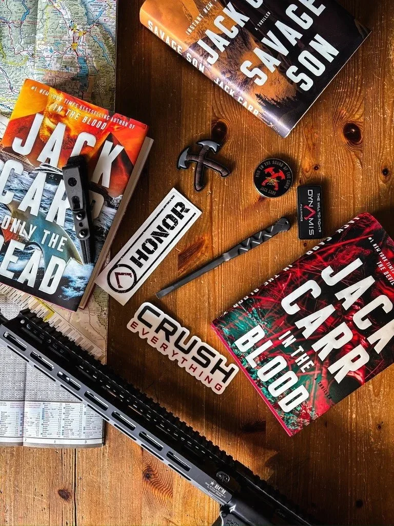 A wooden table with several books by Jack Carr, including "In the Blood," "Only the Dead," and "The Devil's Hand," along with a map, a black tactical knife, a small anchor-shaped item, a round sticker with a dog and red background, a pen, a sticker t