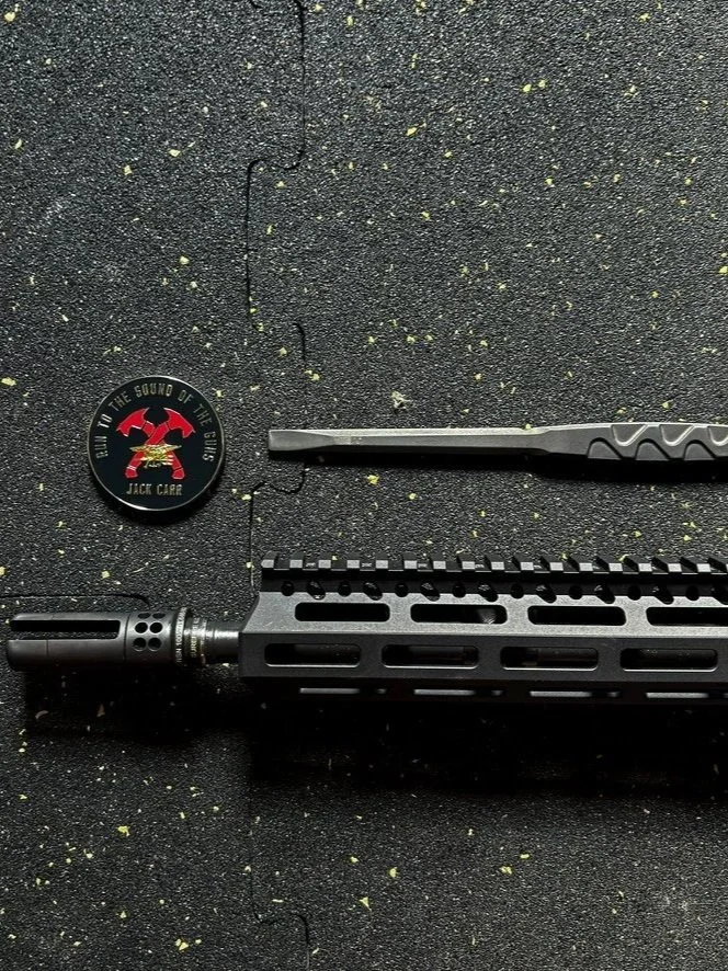 Close-up of a firearm’s handguard, a tactical tool, and a round emblem with a red and black logo and text that reads 'In the Sound of the Guns' by Jack Giar, on a textured black surface with small greenish-yellow specks.