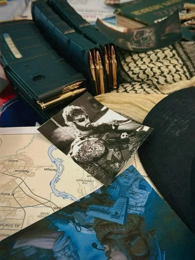 A collection of military and tactical items, including a map, photograph of a soldier with sunglasses holding a gun, and a box of bullets. The scene appears to be a tabletop display.