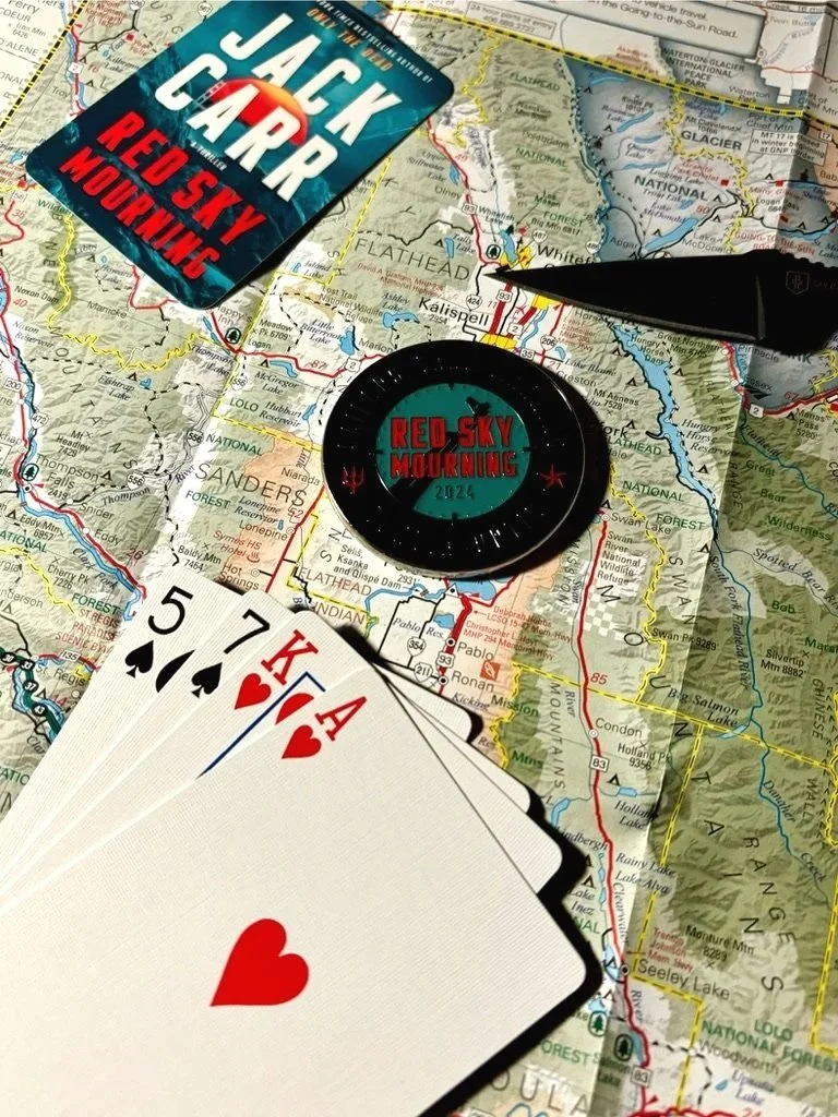 A map with a Jack Carter book titled 'Red Sky Mourning,' a round badge labeled 'Red Sky Mourning 2024,' a closed black pen, and a hand of playing cards showing five, seven, King, Ace, and Ace of hearts, spread over the map.