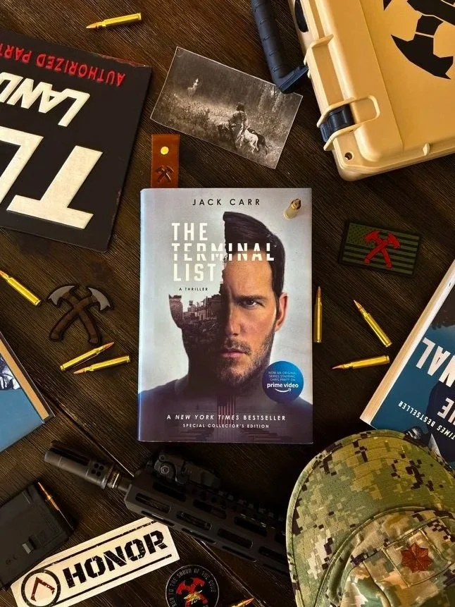 A desk scene featuring a book titled 'The Terminal List' by Jack Carr, surrounded by military-themed items including a camouflage hat, a patch, bullets, a small axe, and other paraphernalia.