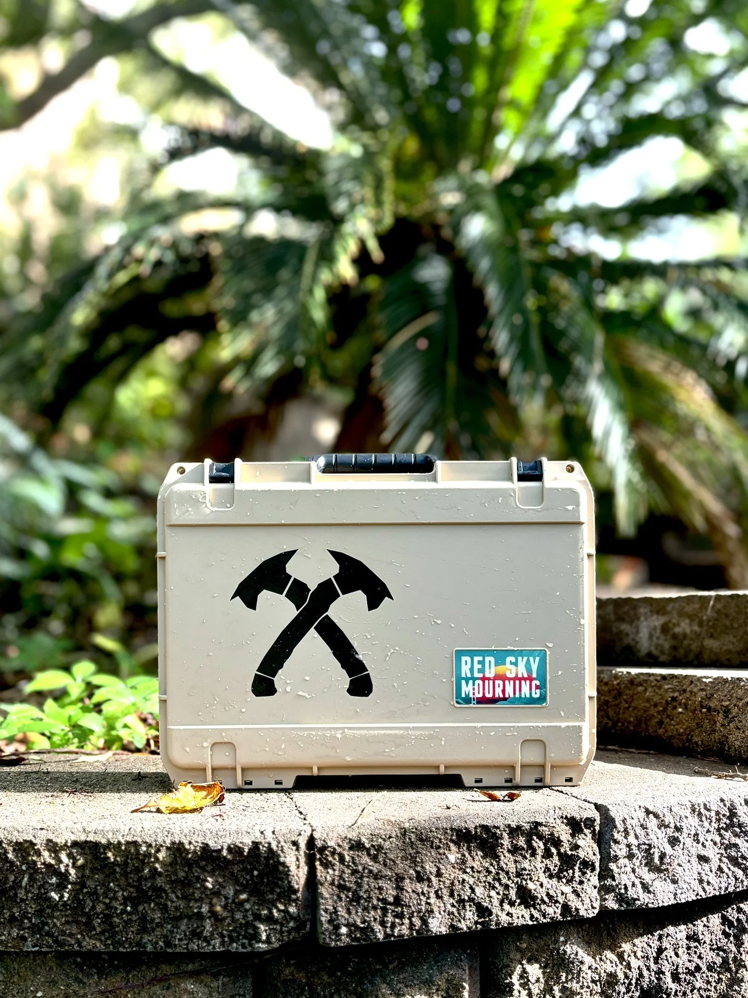 A white box with crossed hammer and axe symbols and a sticker that reads 'Red Sky Mourning' placed on a stone ledge outdoors with greenery and tropical plants in the background.