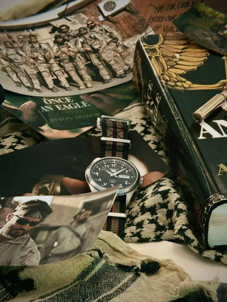 A collection of military-themed items, including a team photo of soldiers in uniform, a book titled "Once an Eagle," a Seiko wristwatch with a striped NATO strap, a photograph of a soldier, and a decorative box with eagle feathers.