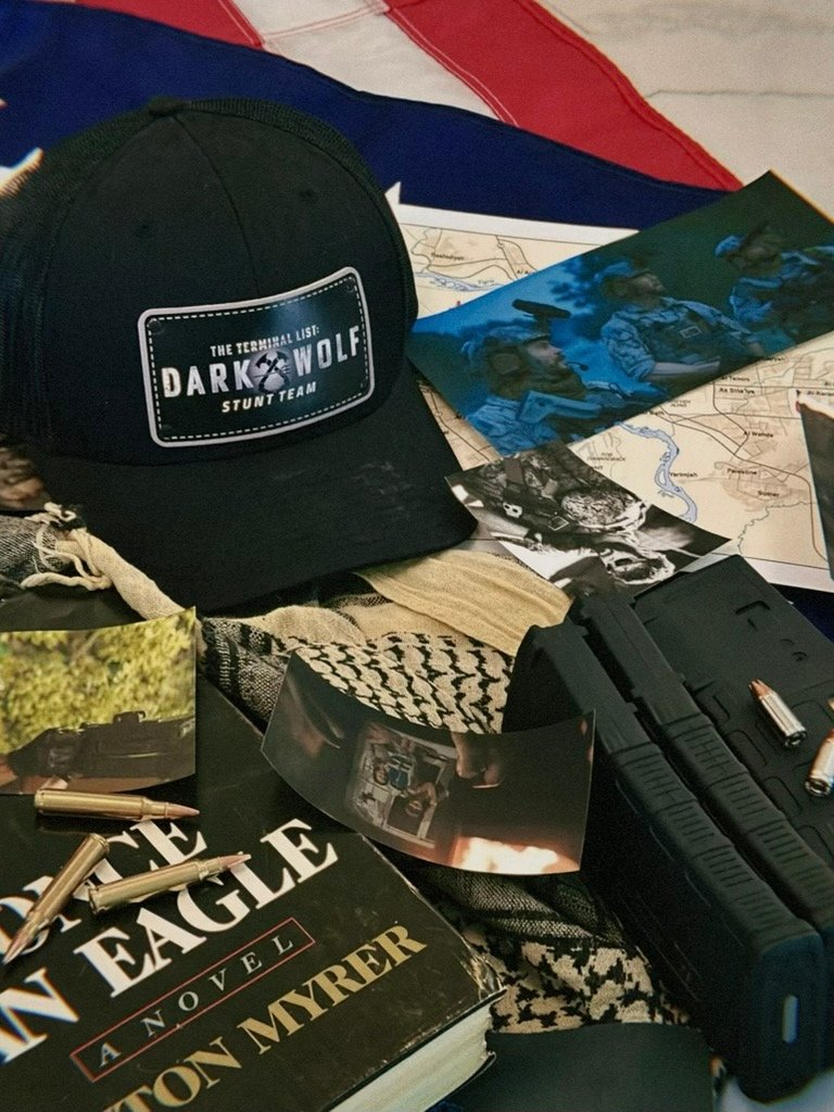 A black cap with a patch that reads 'The Terminal List: Dark Wolf Stunt Team' is placed among books, photos, a bag of ammunition, and maps, suggesting a military or tactical theme.