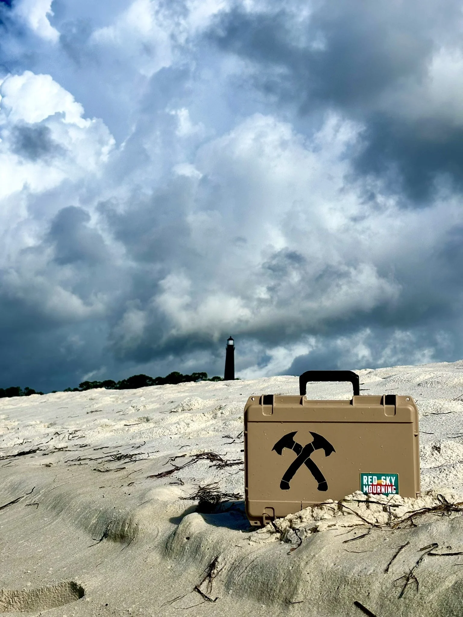 A beige toolbox with crossed hammers logo and a sticker reading "Red Sky Mourn" on a sandy beach with a lighthouse in the distance and stormy clouds overhead.