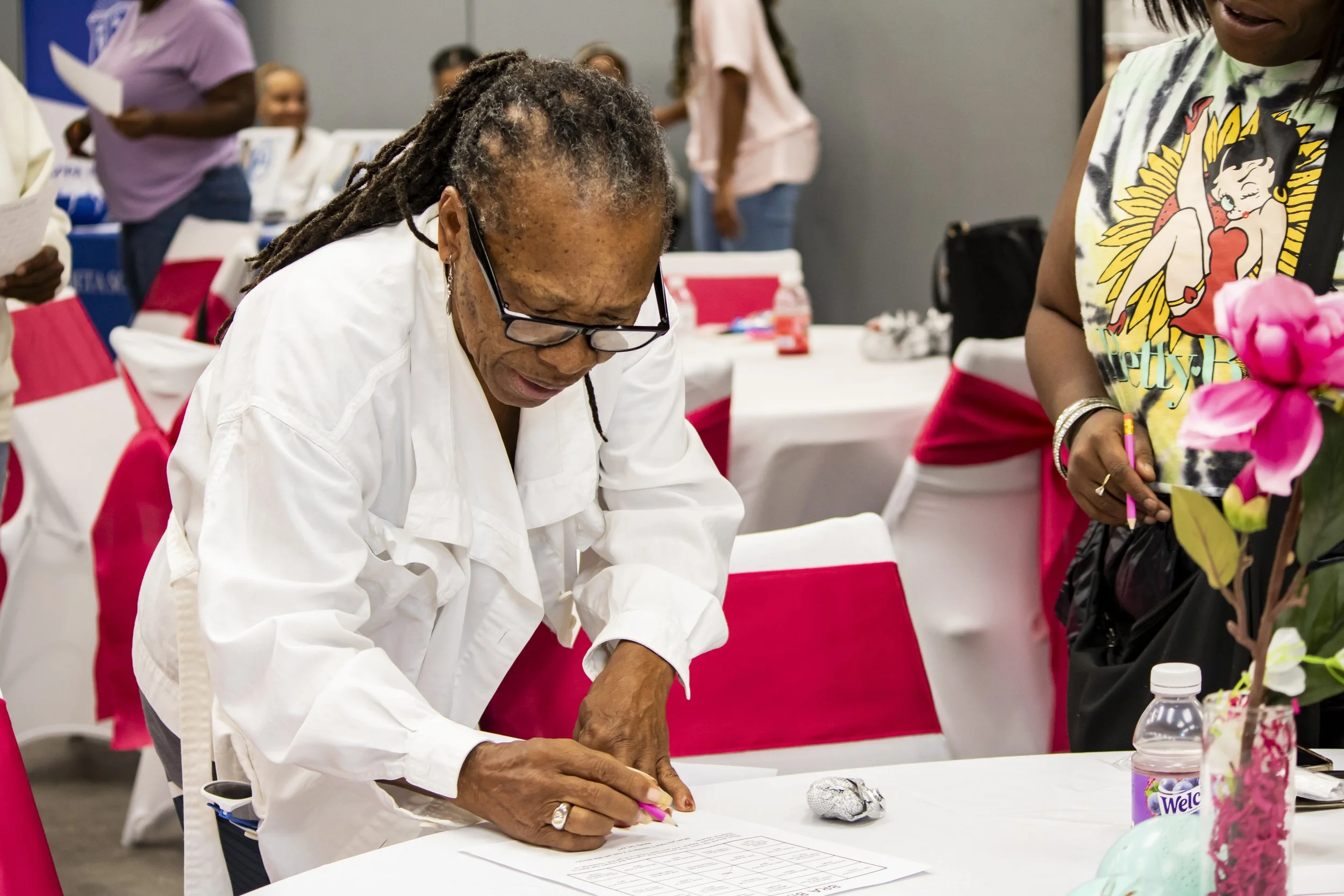 An elderly woman in a white coat and glasses signing a document at a table with pink decorations during an indoor event.