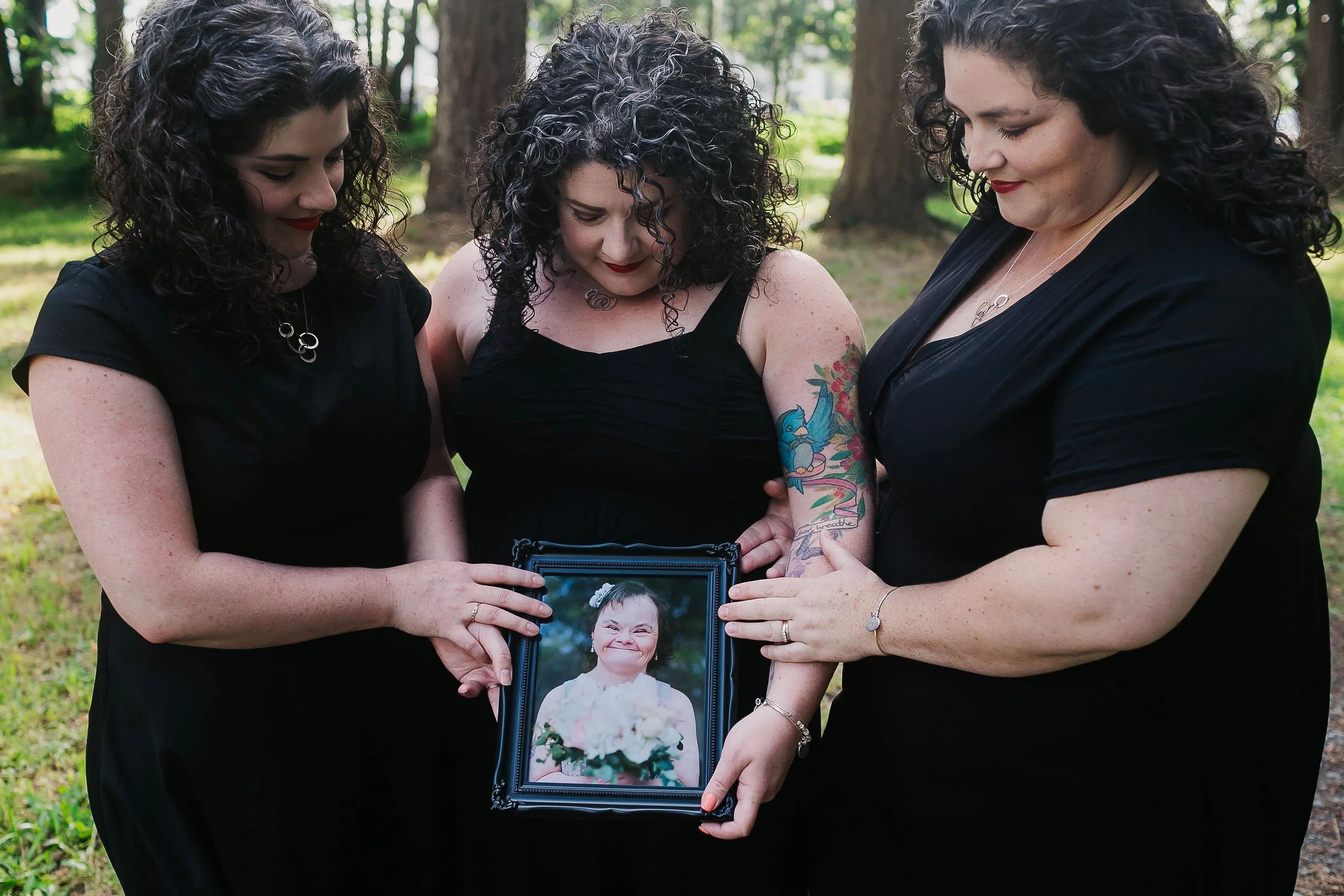 Three women in black dresses holding a framed photograph of a woman with a big smile, outdoors in a wooded area.