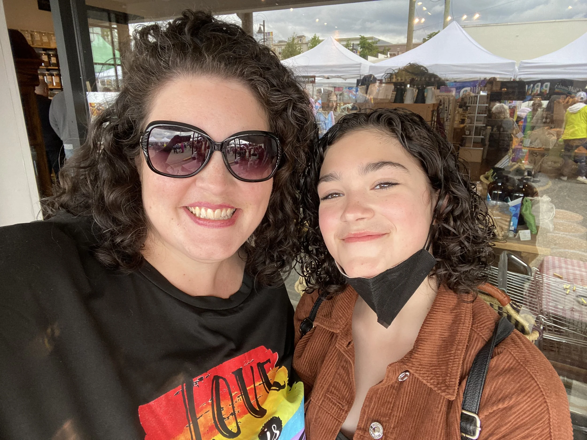 Two smiling women, one wearing sunglasses, taking a selfie outside at a market or festival with tents and people in the background.