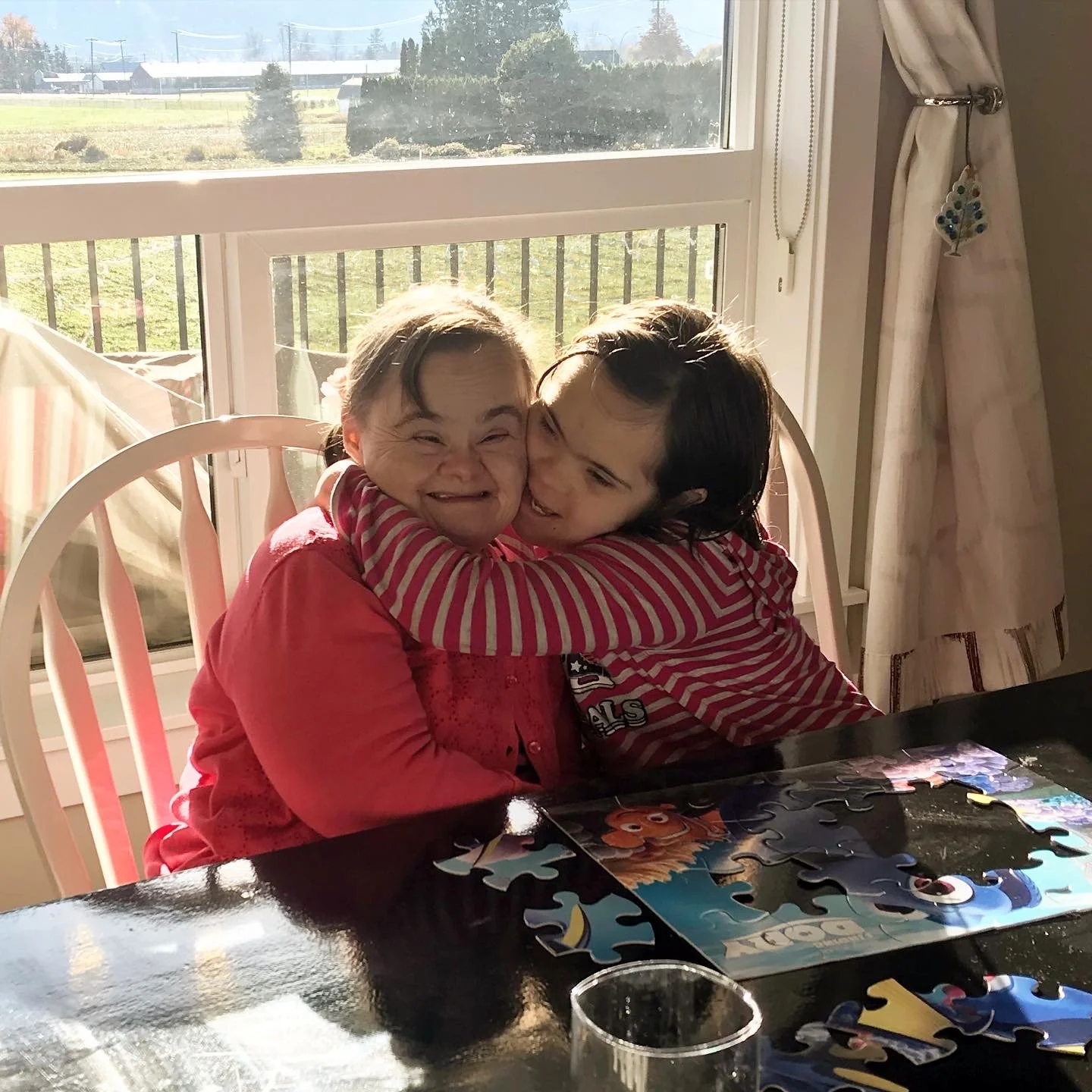 Two children are hugging and smiling at a table with a partially completed puzzle in front of a window with a scenic view outside.