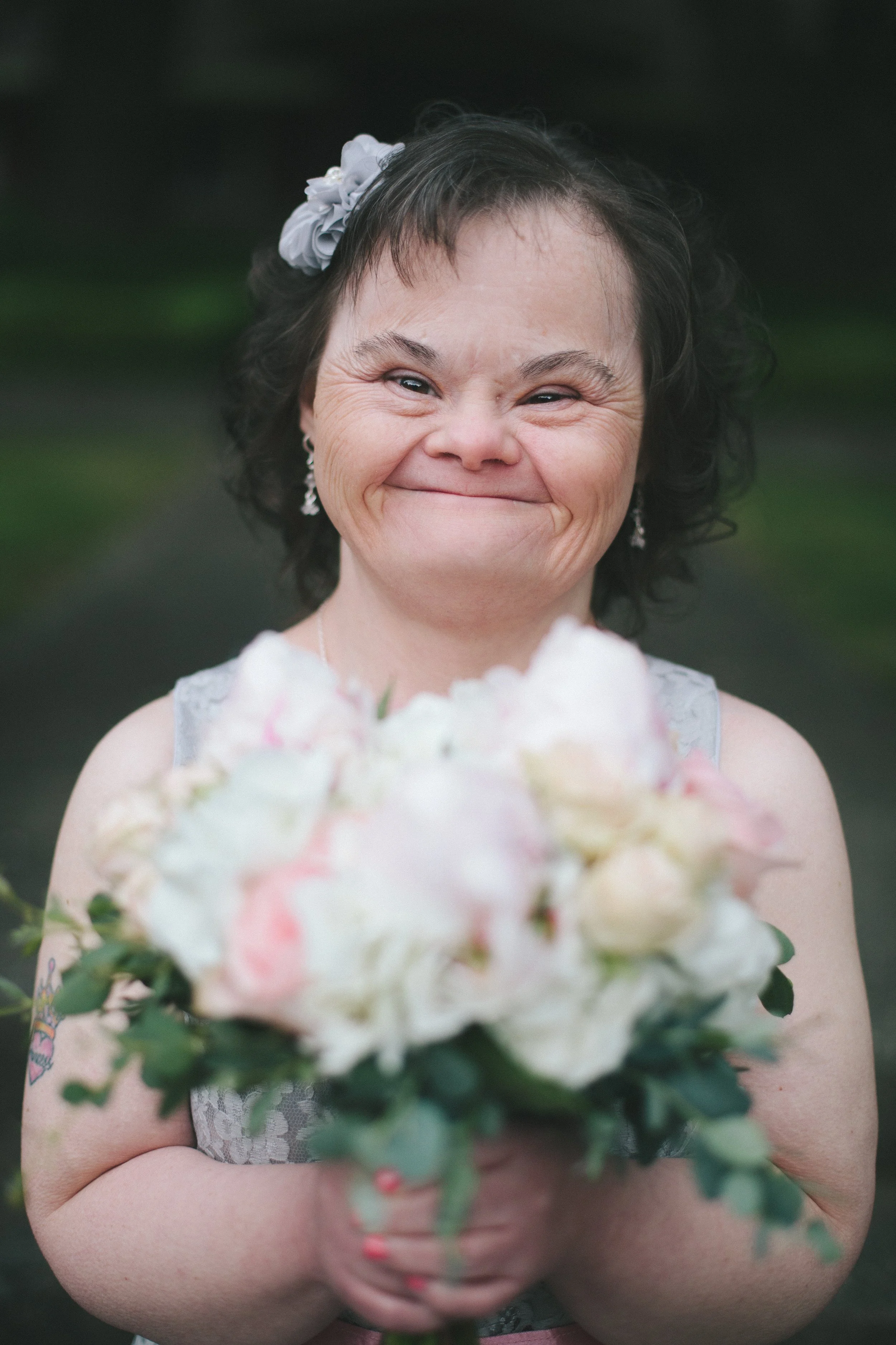A woman with short dark curly hair, wearing a light-colored dress, smiling and holding a bouquet of pastel-colored flowers, outdoors with a blurred green background.