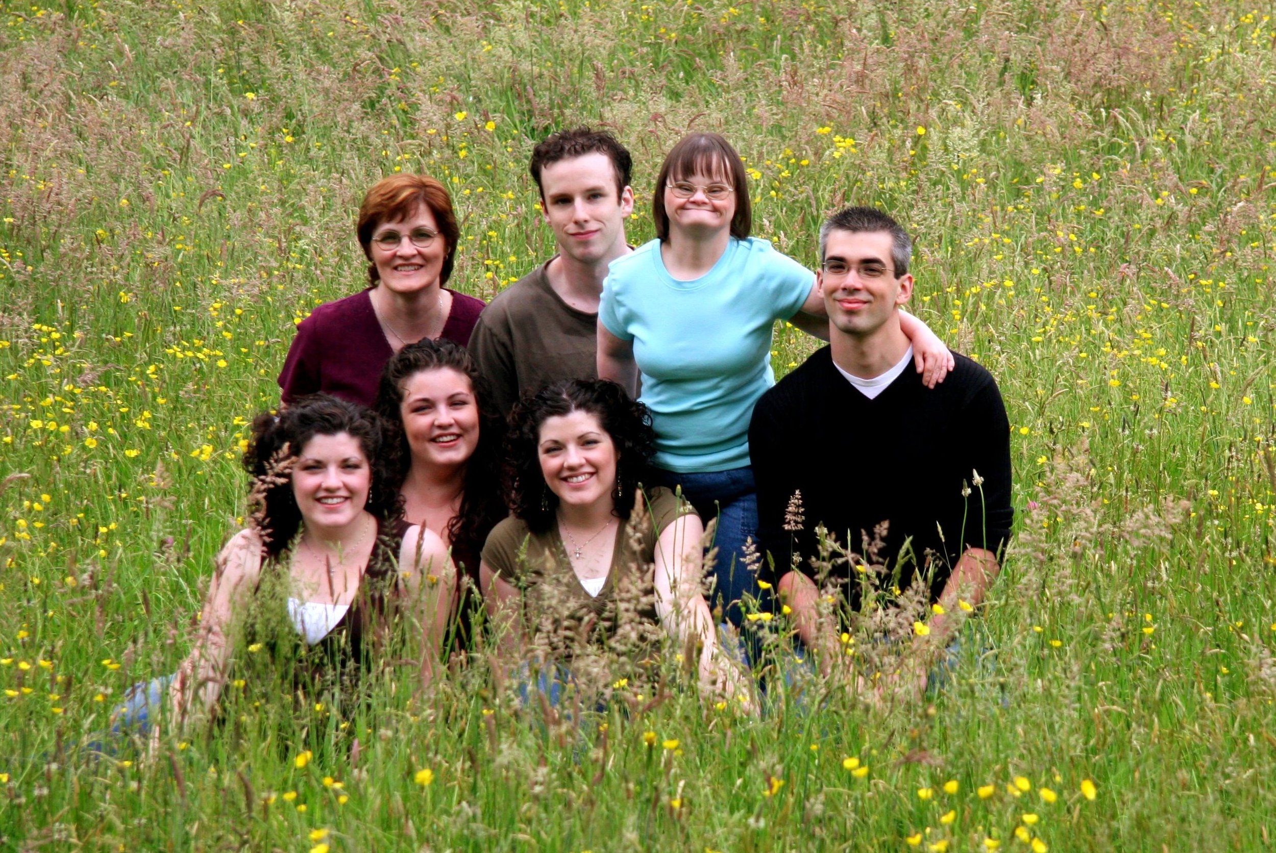 A family of eight people, including three women, three men, and two young women, sitting and standing in a grassy field with yellow flowers, smiling at the camera.