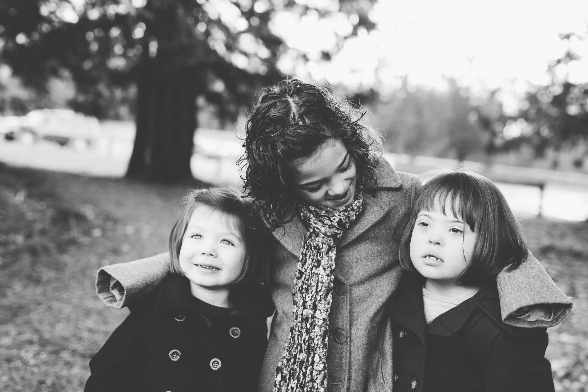 A woman with curly hair is smiling and looking down at two young girls, one on each side, outdoors in a park.