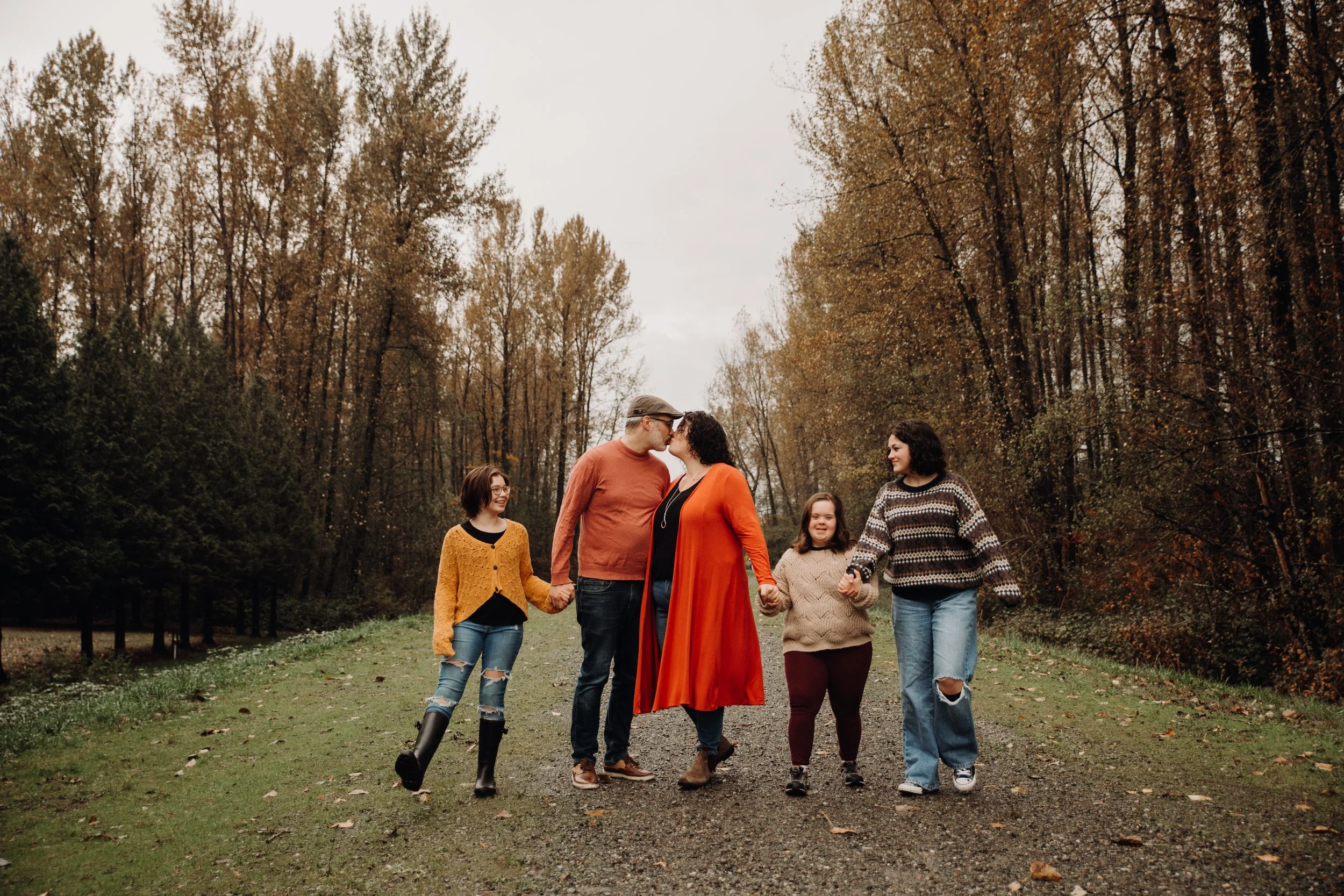 A family of six walking hand-in-hand outdoors on a forest path in autumn, surrounded by tall trees with fall foliage.
