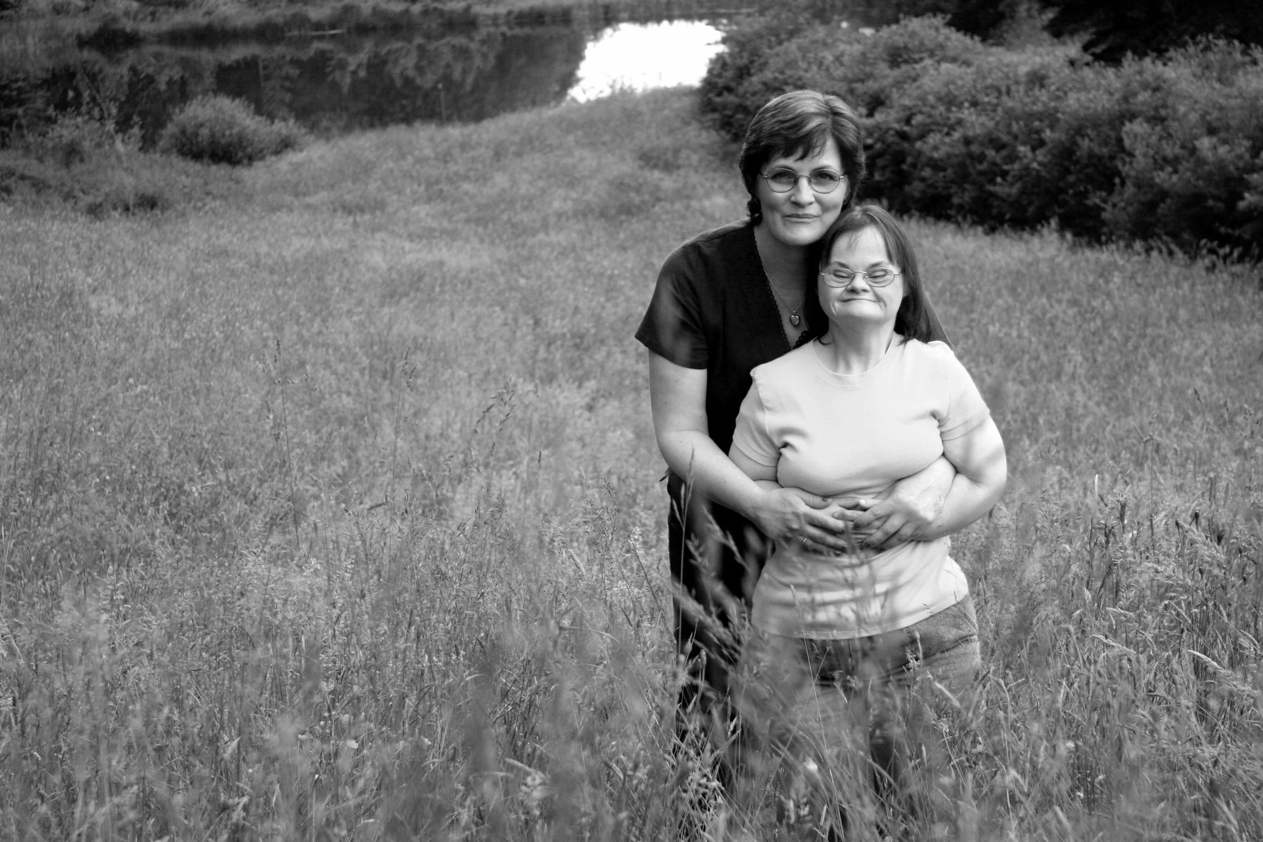 A woman and a girl standing together in a grassy field, smiling at the camera, with trees and a pathway in the background.