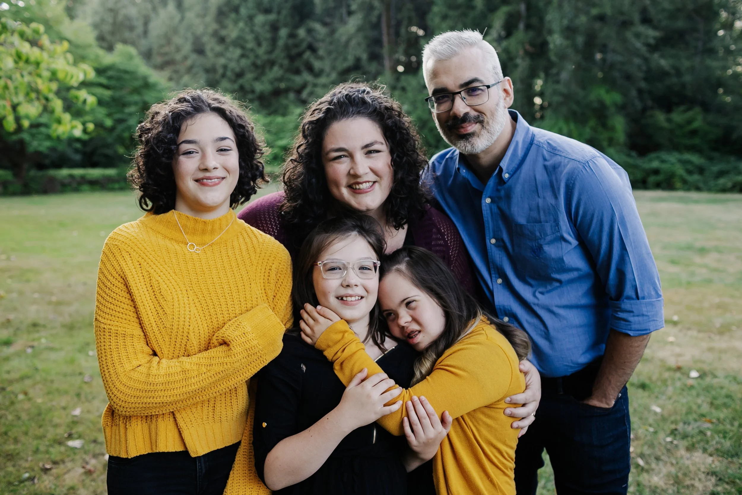 A multigenerational family of six posing outdoors in a park, smiling and hugging each other. The group includes two adults, two teenagers, and two children, all wearing colorful casual clothing. There are green trees and grass in the background.