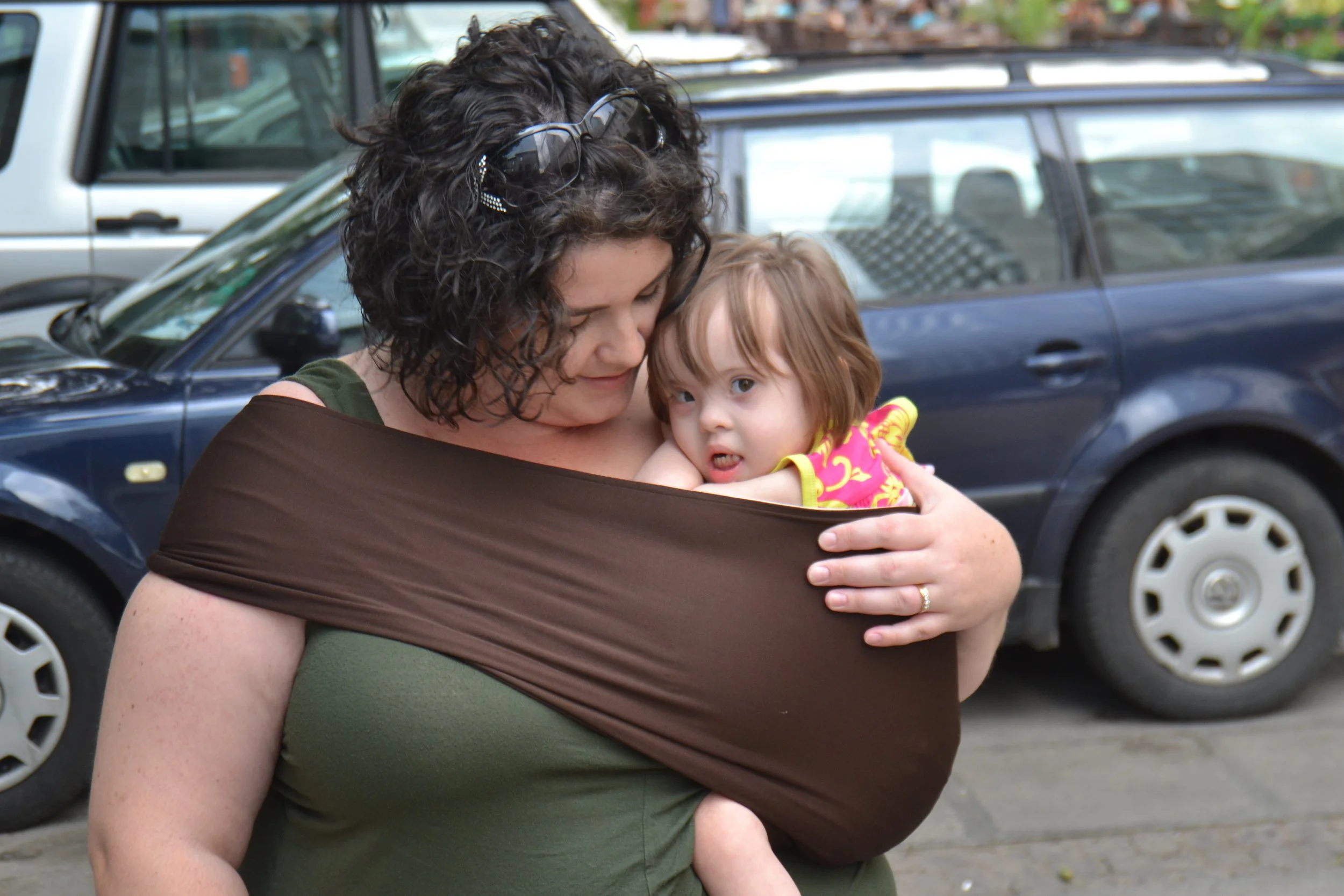 A woman with curly dark hair and sunglasses on her head holds a young girl in a colorful dress, both cuddling outside near parked cars.