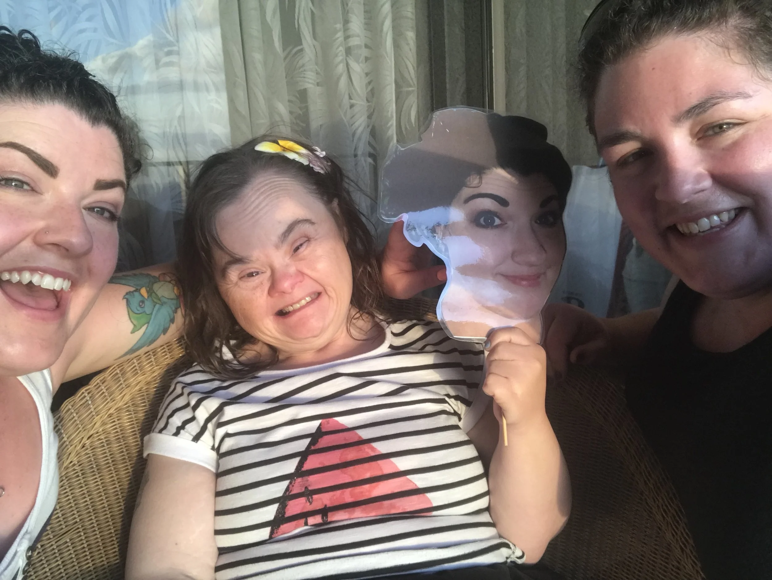 Four smiling people taking a selfie, with one holding a mask of a woman's face, sitting on a wicker chair near a window with curtains.