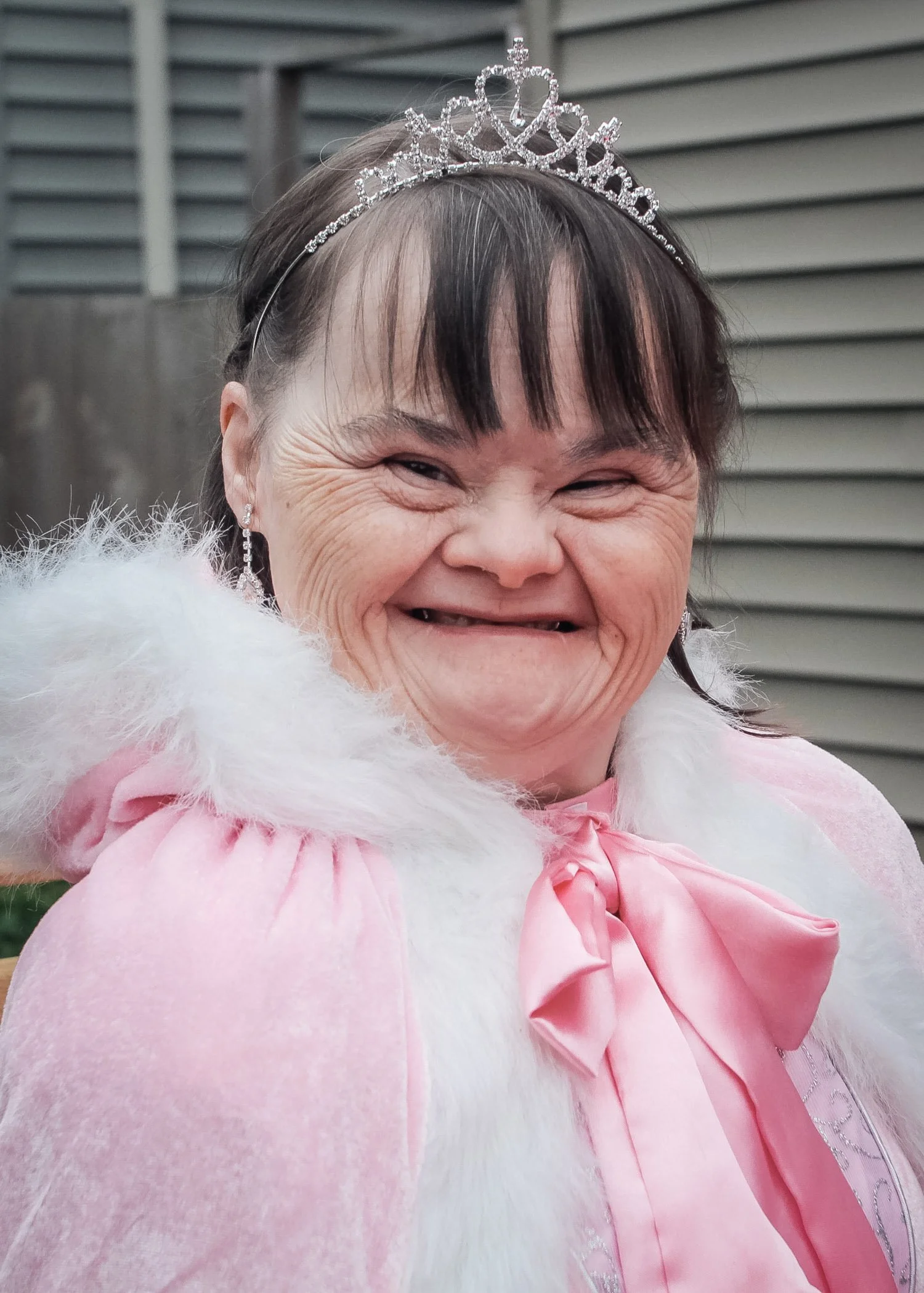 A smiling woman dressed as a princess, wearing a pink dress with a pink bow and a white feathered cape, with a tiara on her head and jewelry, outdoors near a house with siding.