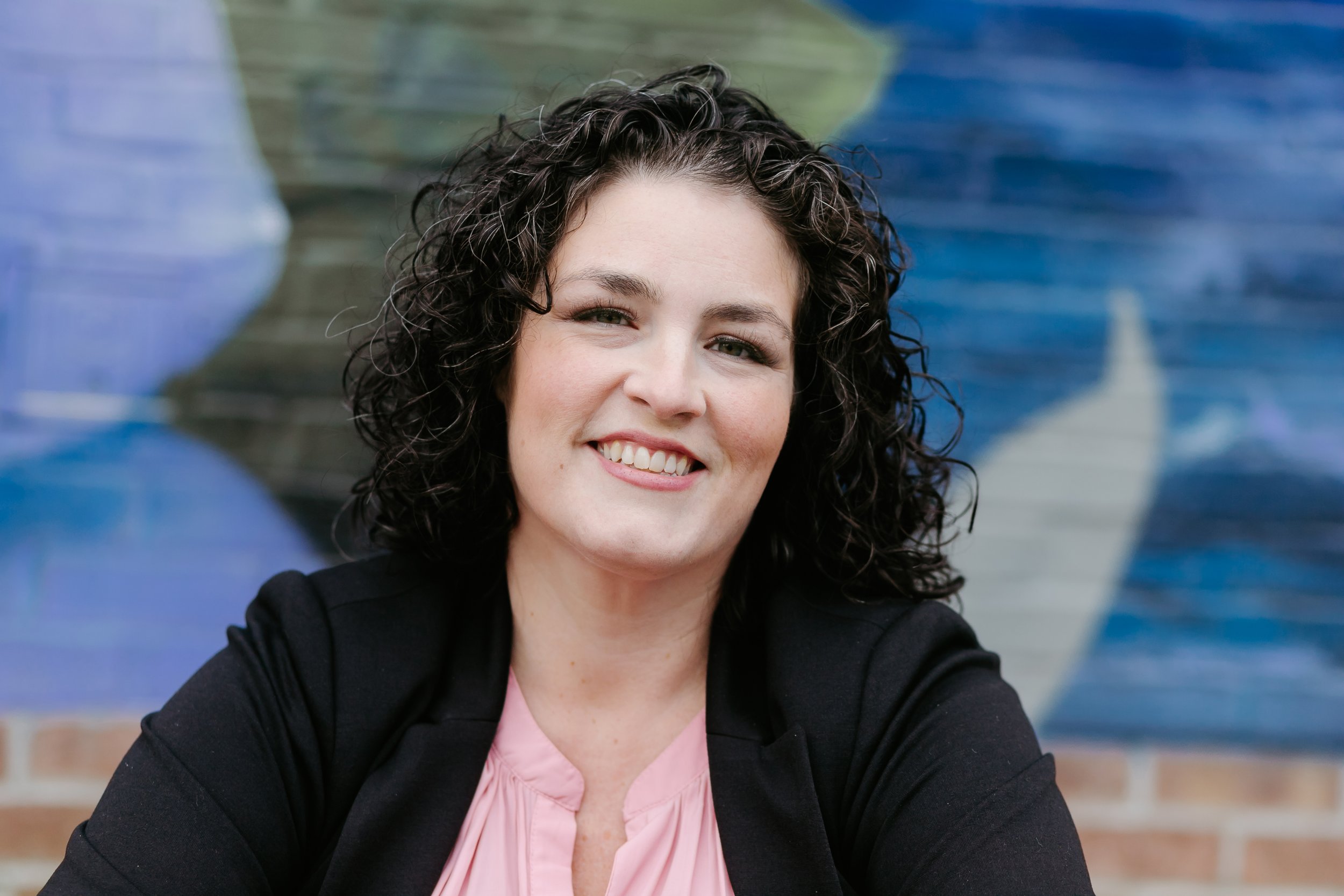 A woman with curly dark hair smiling, wearing a black blazer and pink blouse, with colorful abstract painting in the background.