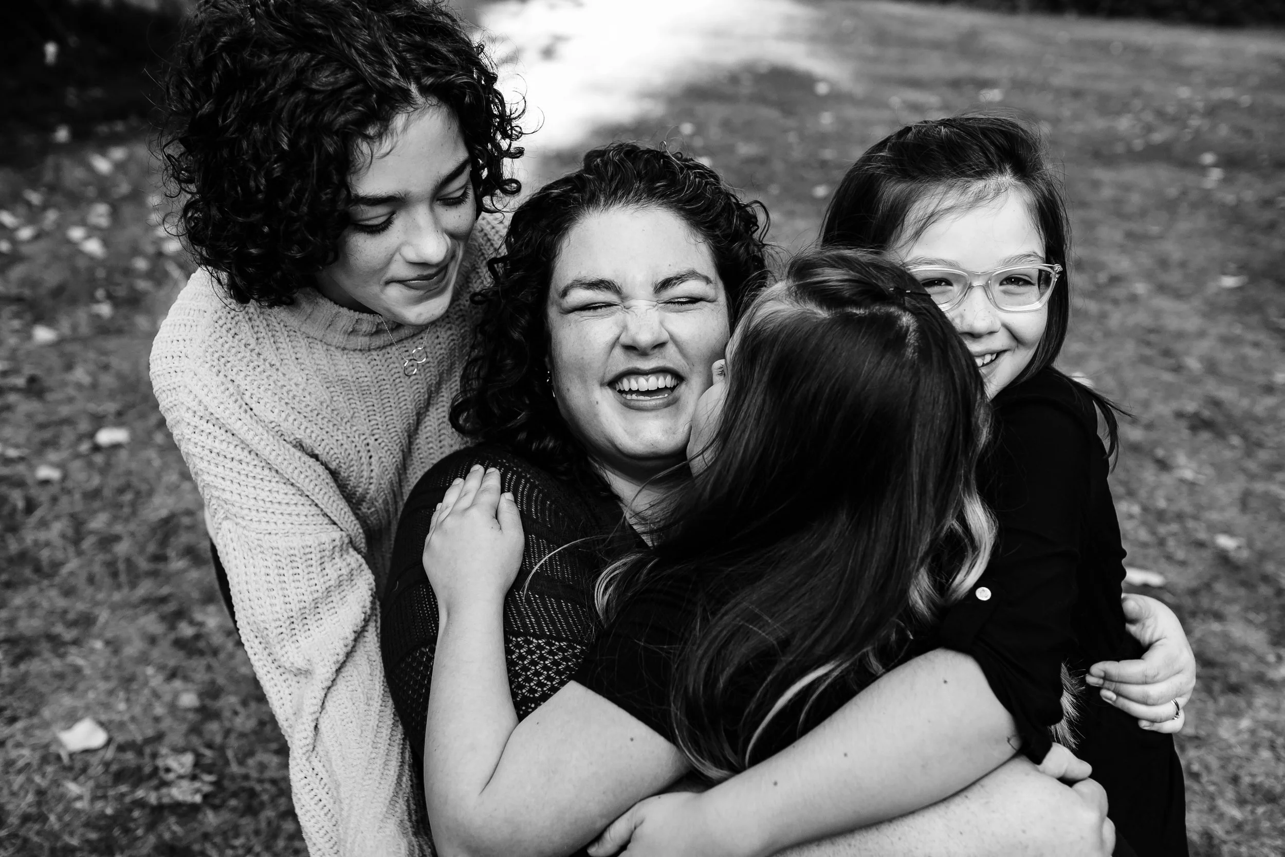 Group of five women hugging and smiling outdoors on a grassy area.