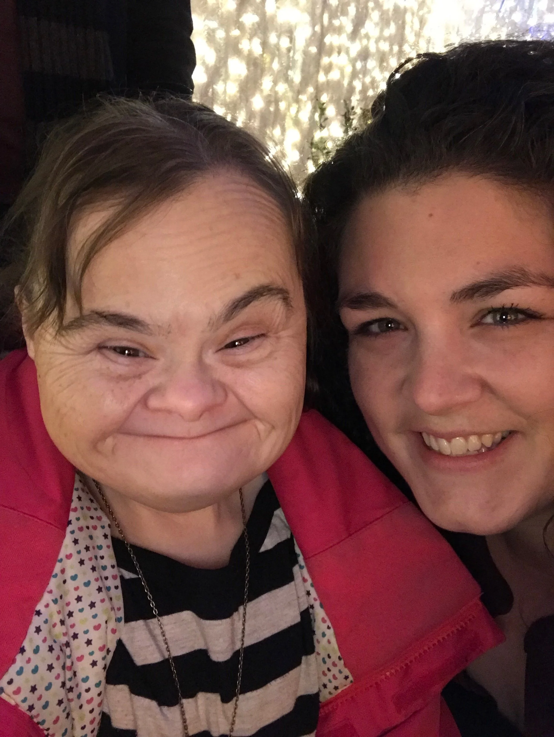 Two smiling women taking a close-up selfie together, outdoors with a background of sunlight filtering through trees.