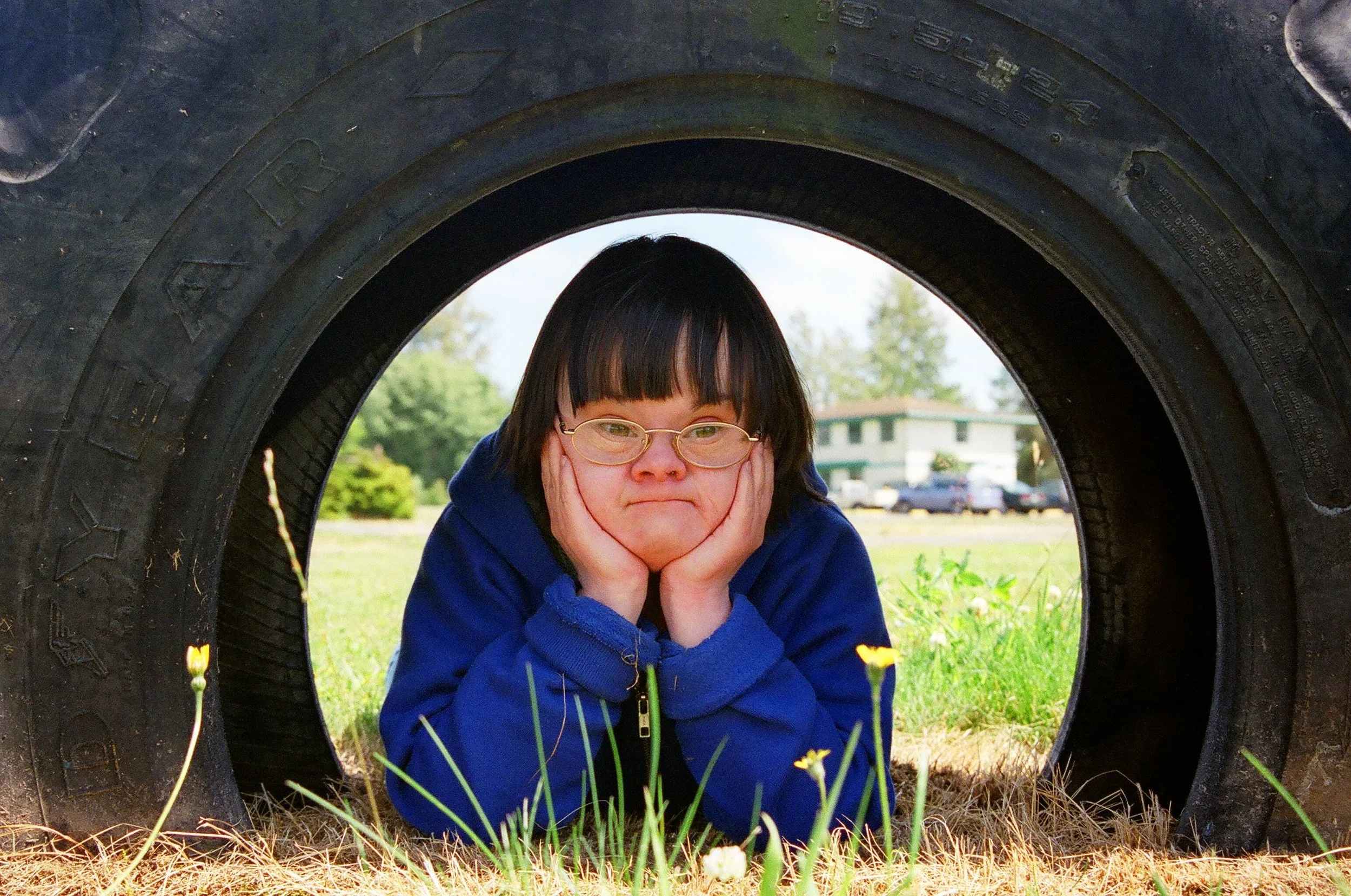 Child with glasses laying on grass, resting their chin on their hands, inside a large tire outdoors.